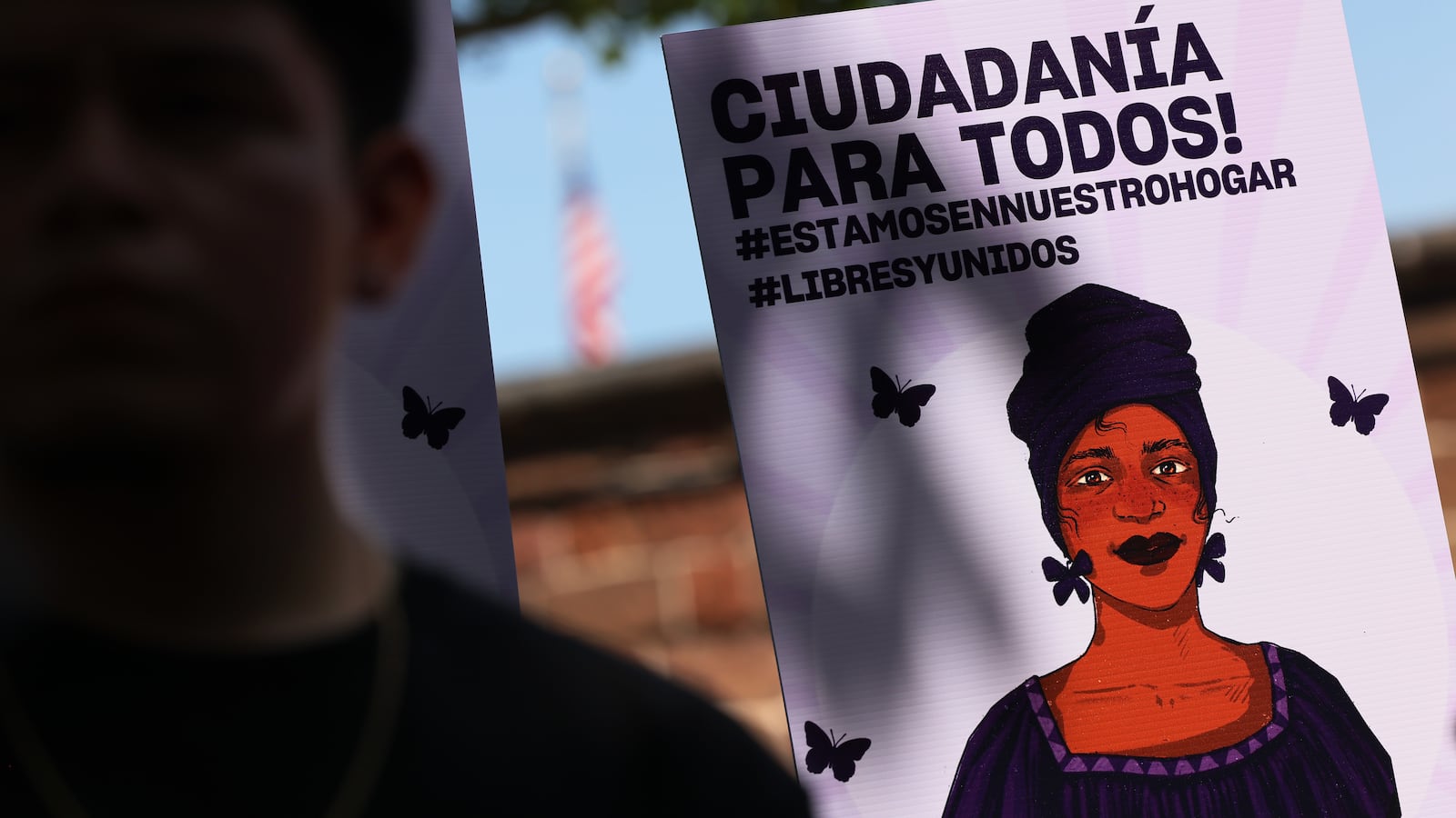 People gathered with one holding up a sign featuring the image of a woman. It reads "Ciudadania Para Todos."