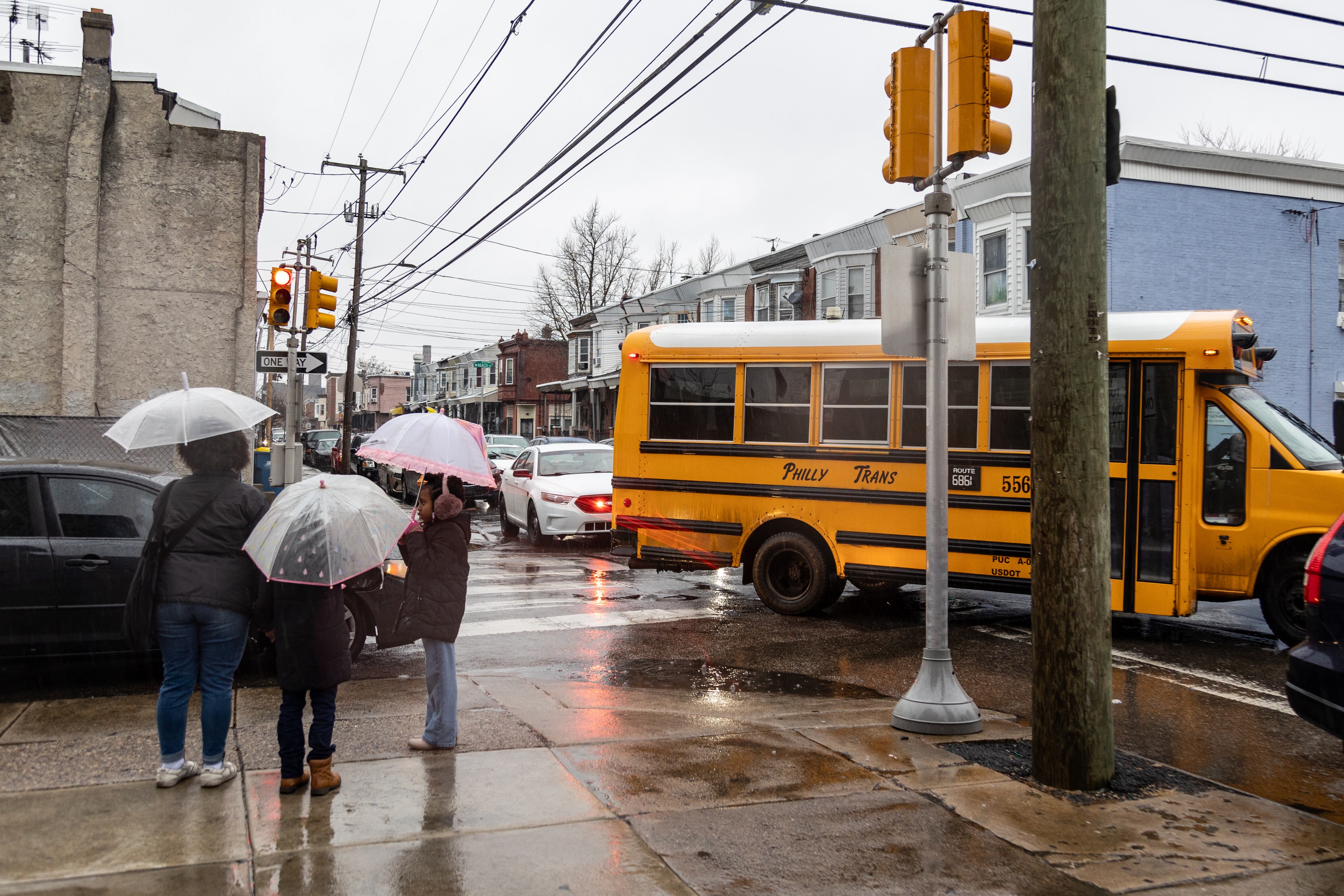 Three people holding umbrellas stand outside at a cross street with a yellow school bus in the backgrond.