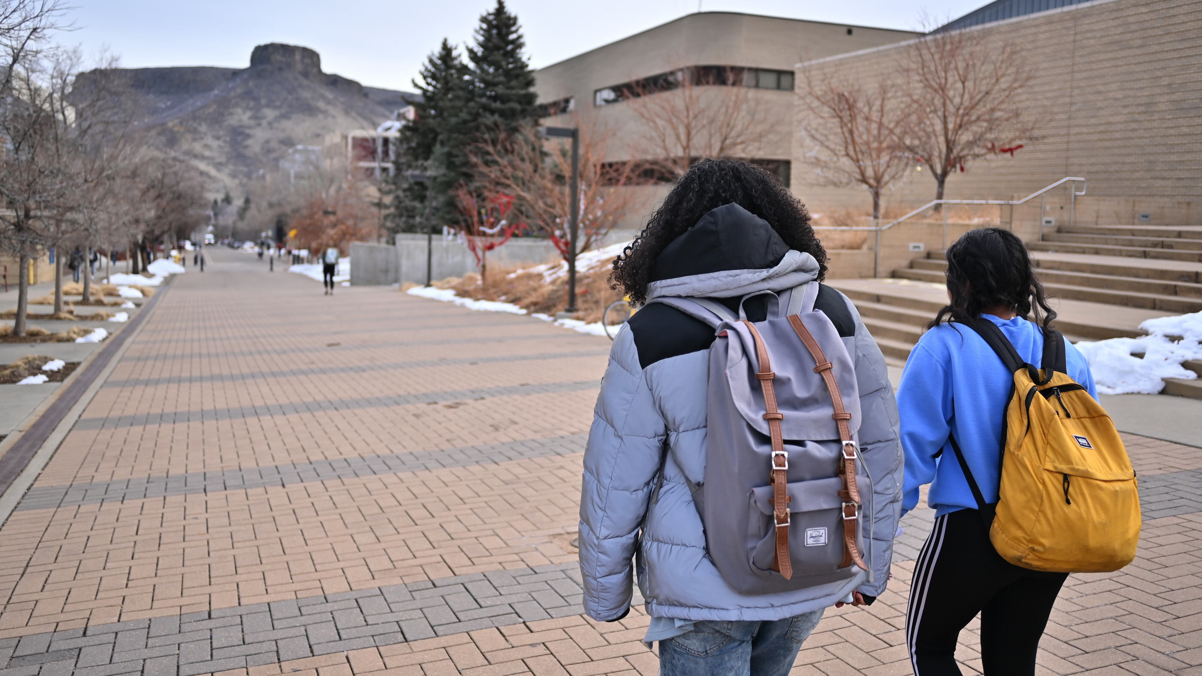 Two students with backpacks on walk on on a college campus with a mountain in the background.