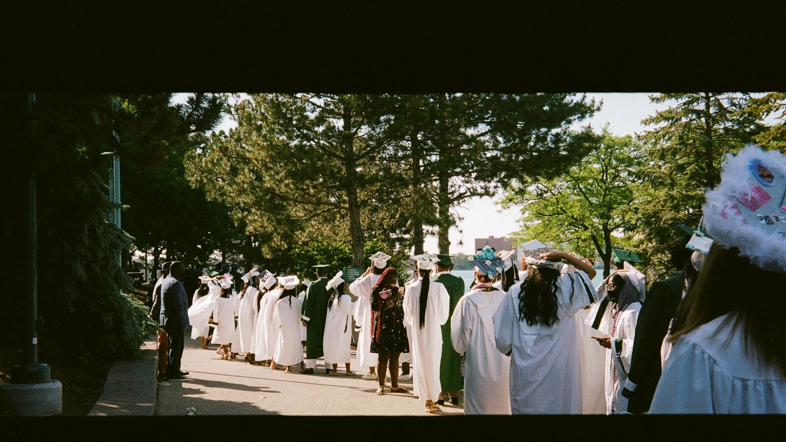 A long line of students in green and white graduation regalia walk through green trees to their ceremony.