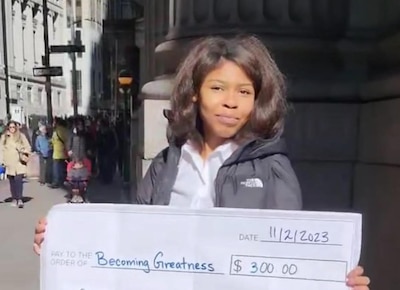 A young woman holds a giant check addressed for $300 to "Becoming Greatness."