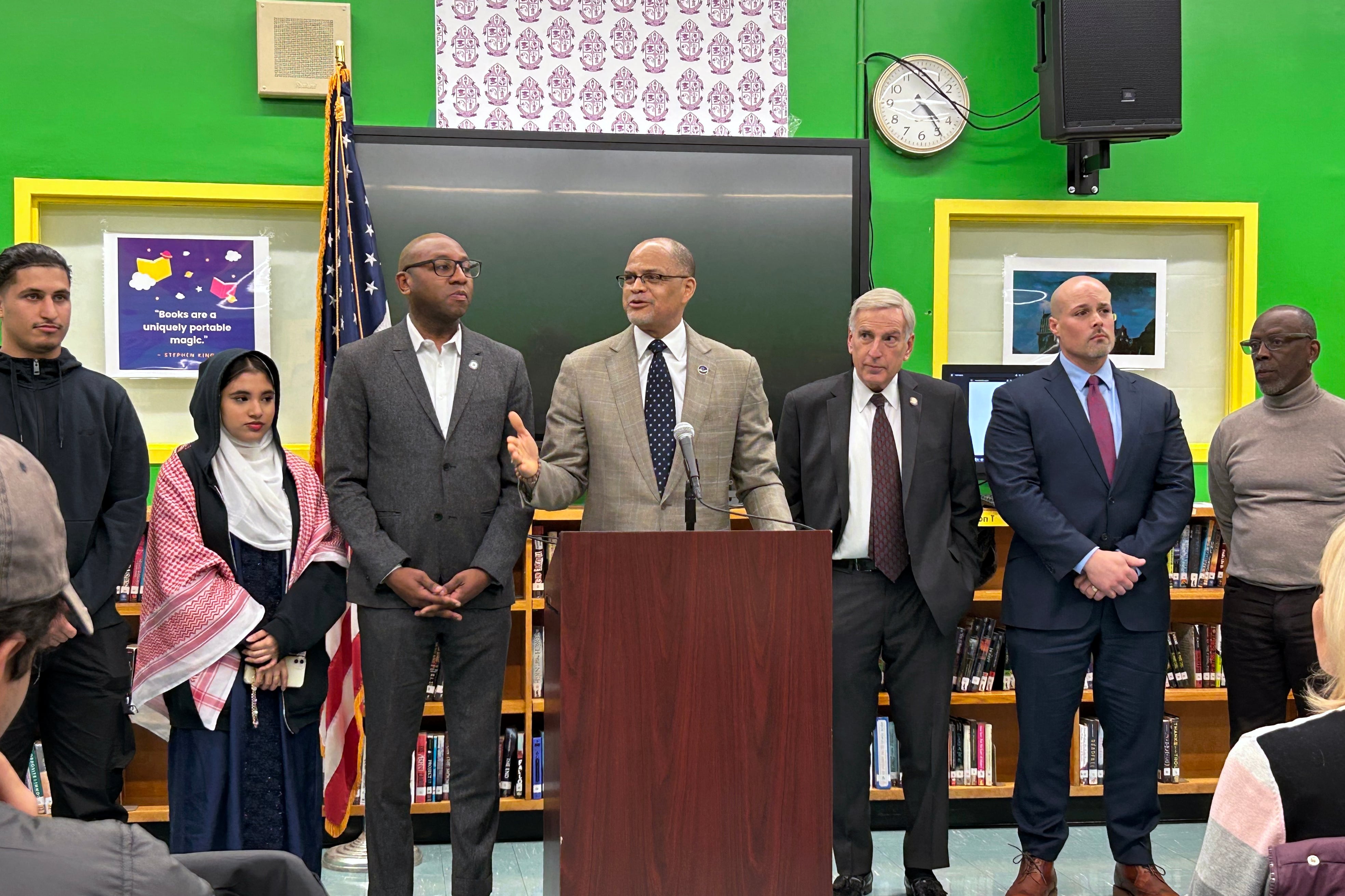 Seven people stand side by side while the person in the middle speaks at a podium. There is a large, black, computer screen, a clock and some posters on the wall in the background.