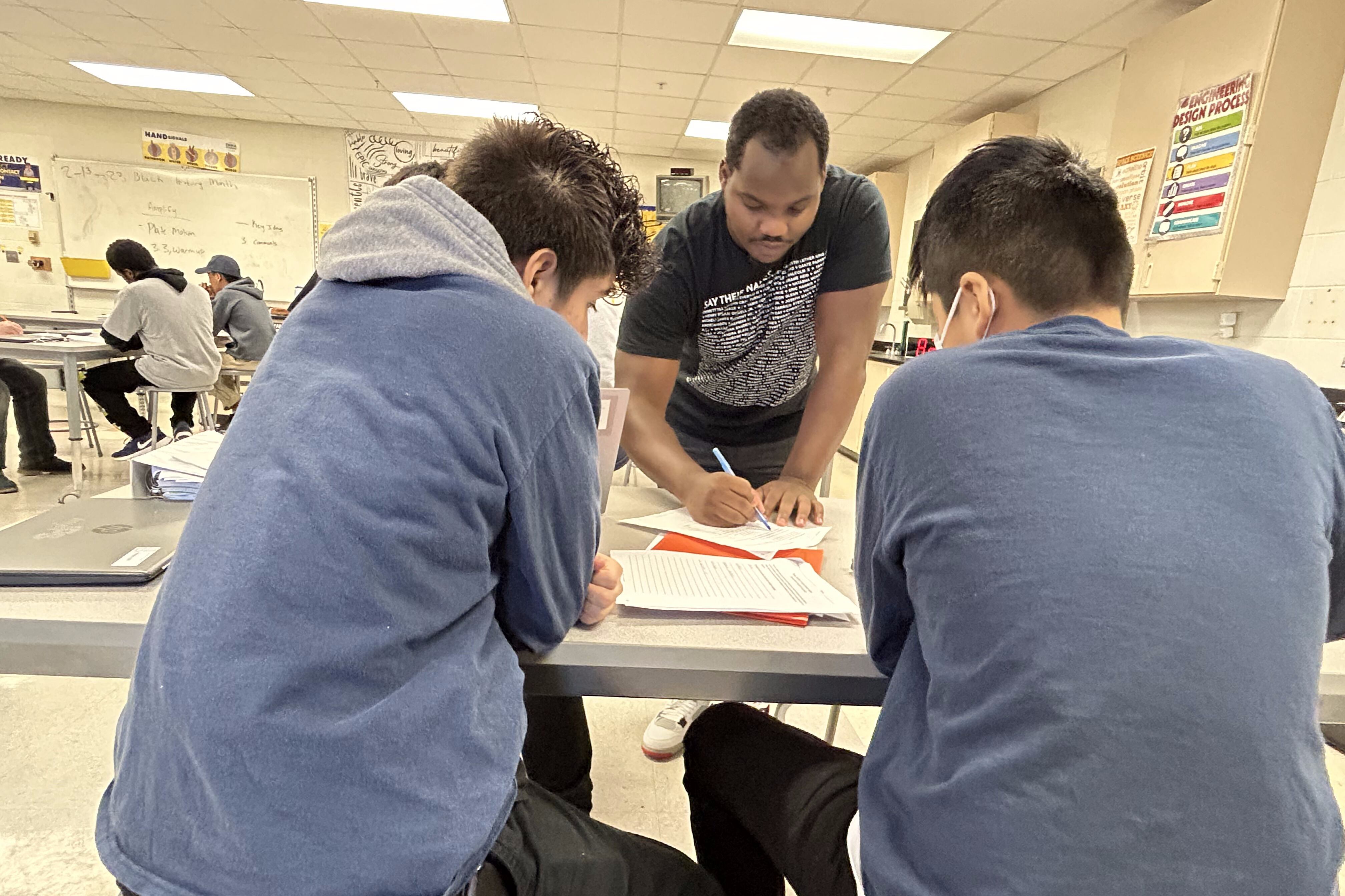 A man stands over two students sitting down at a table, reviewing their school work with them.