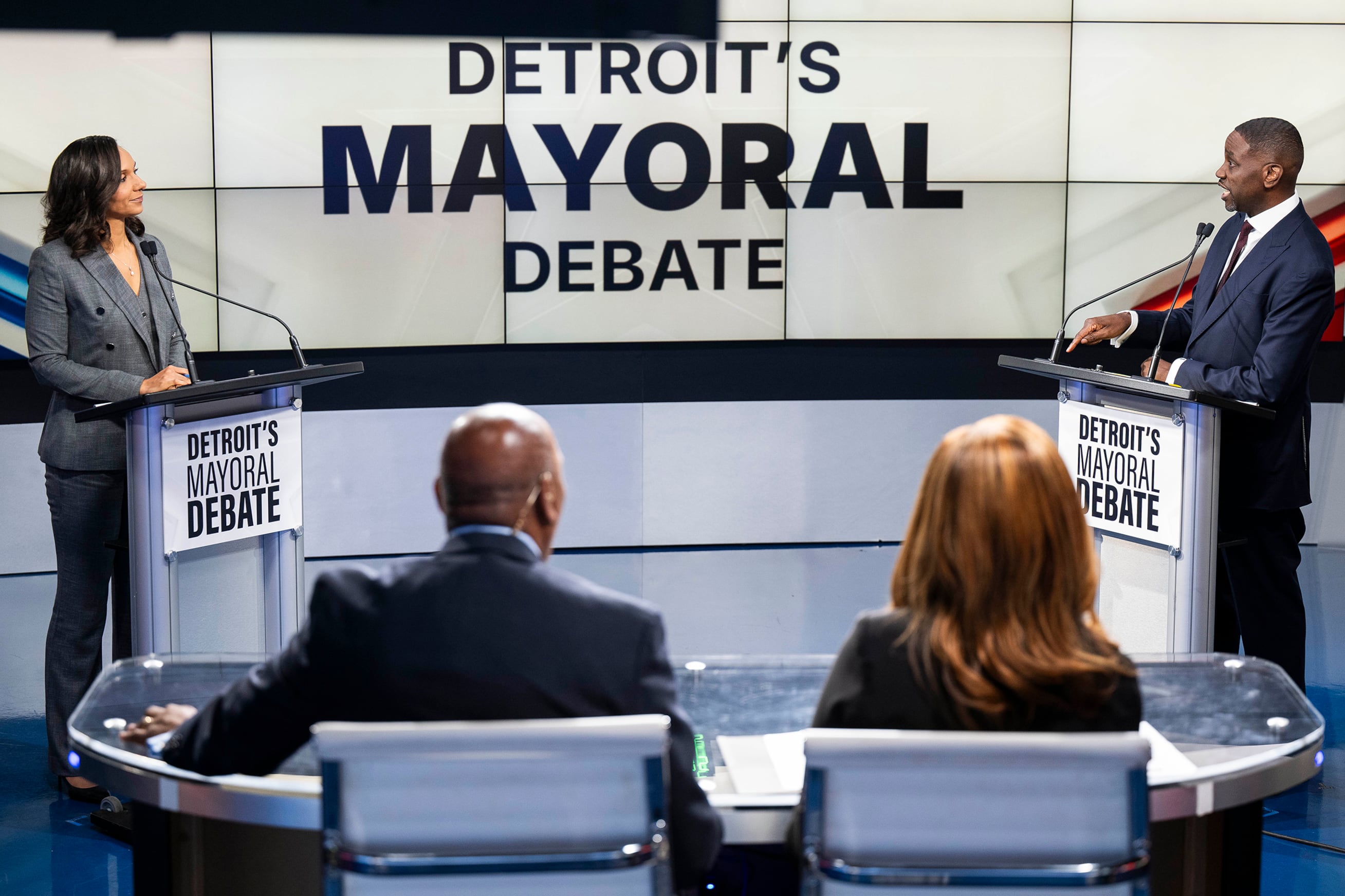 A photograph of two adults in suits standing behind podiums during a debate with two people sitting in the foreground.