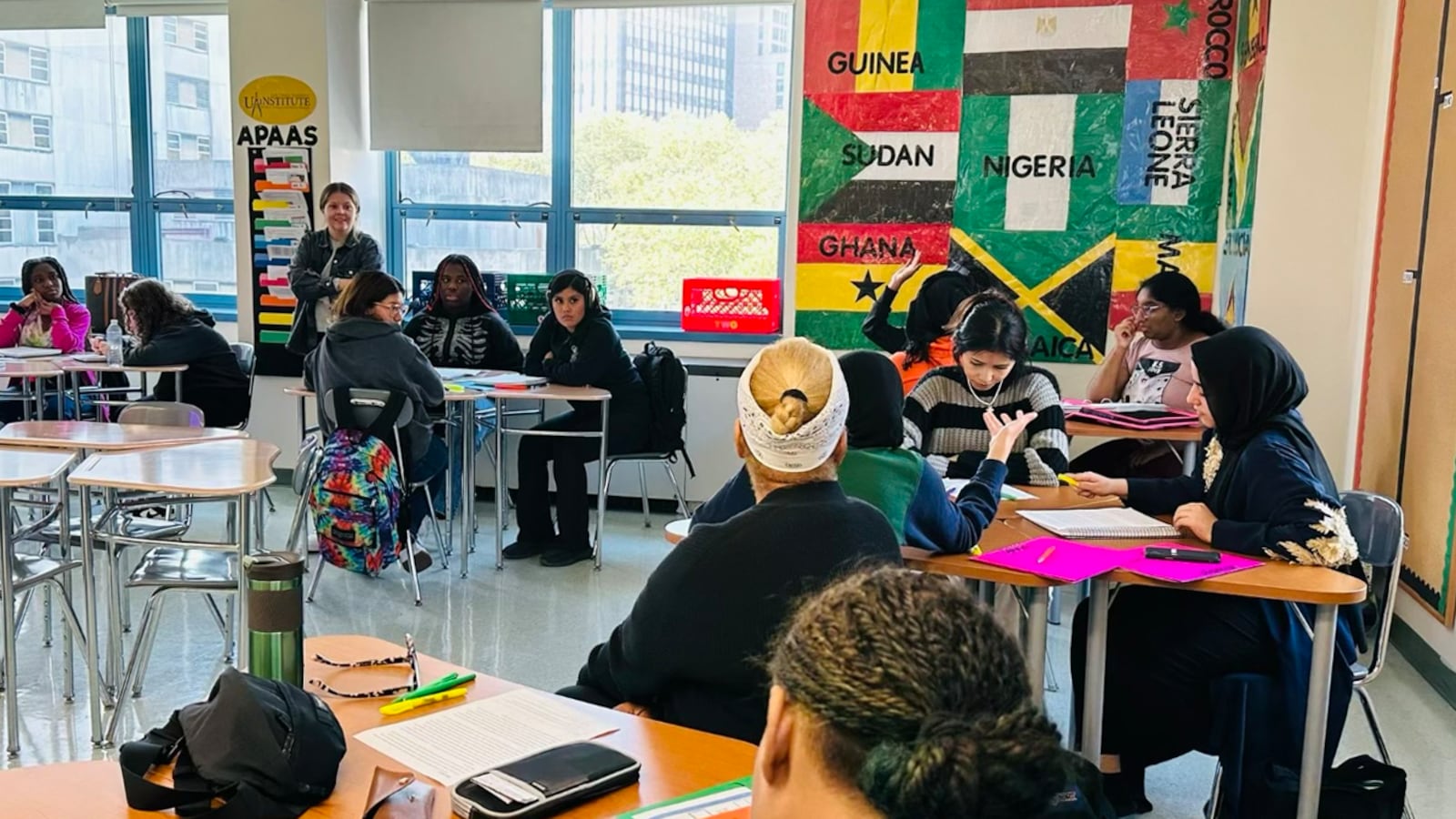 Students sit at desks in a classroom with colorful flags on the back wall next to large windows.