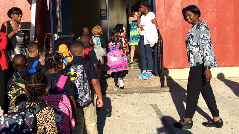 A line of students wearing backpacks walks into an elementary school. 
