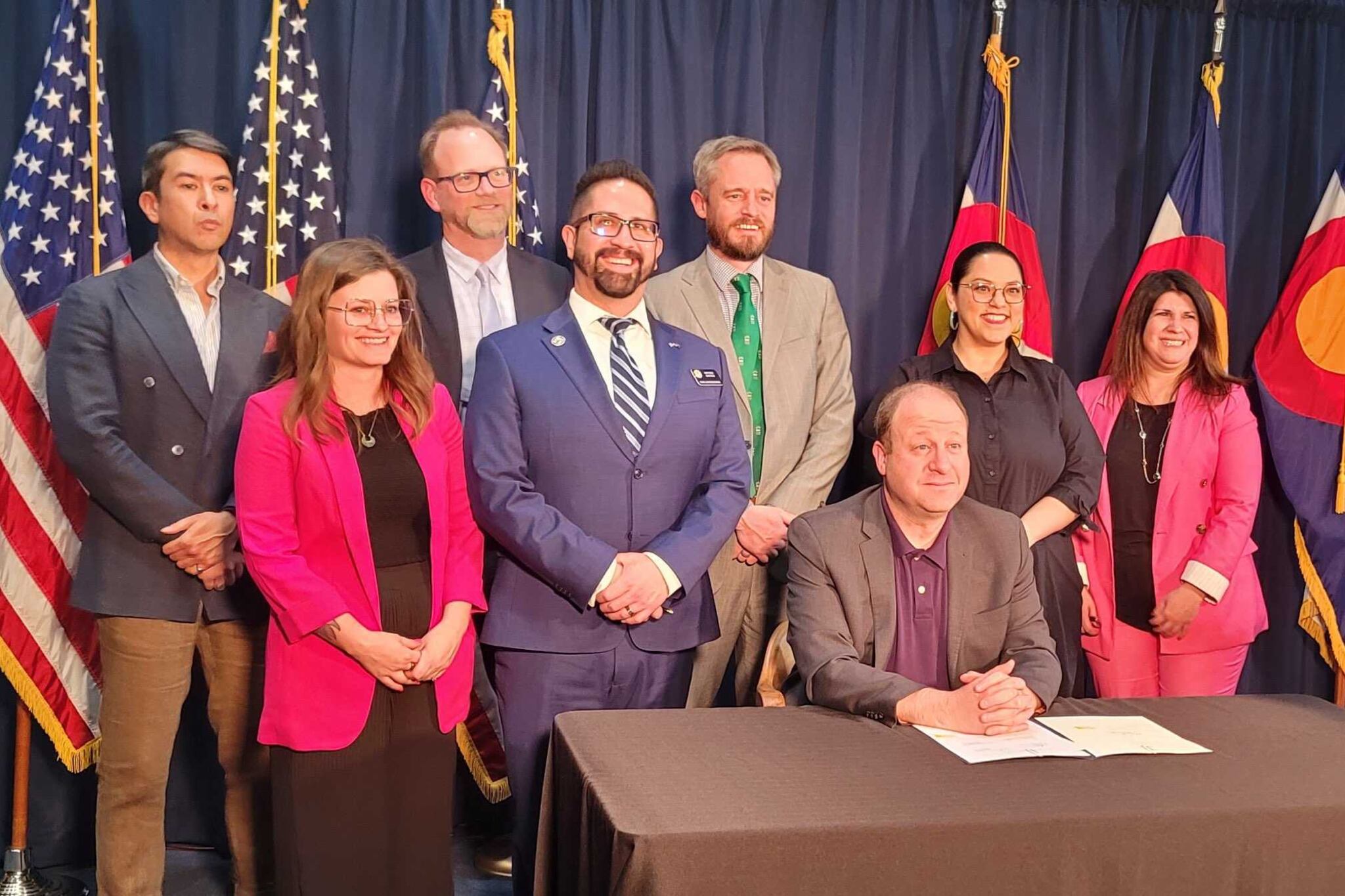 Colorado lawmakers stand together in front of a blue background and Colorado and United States flags.