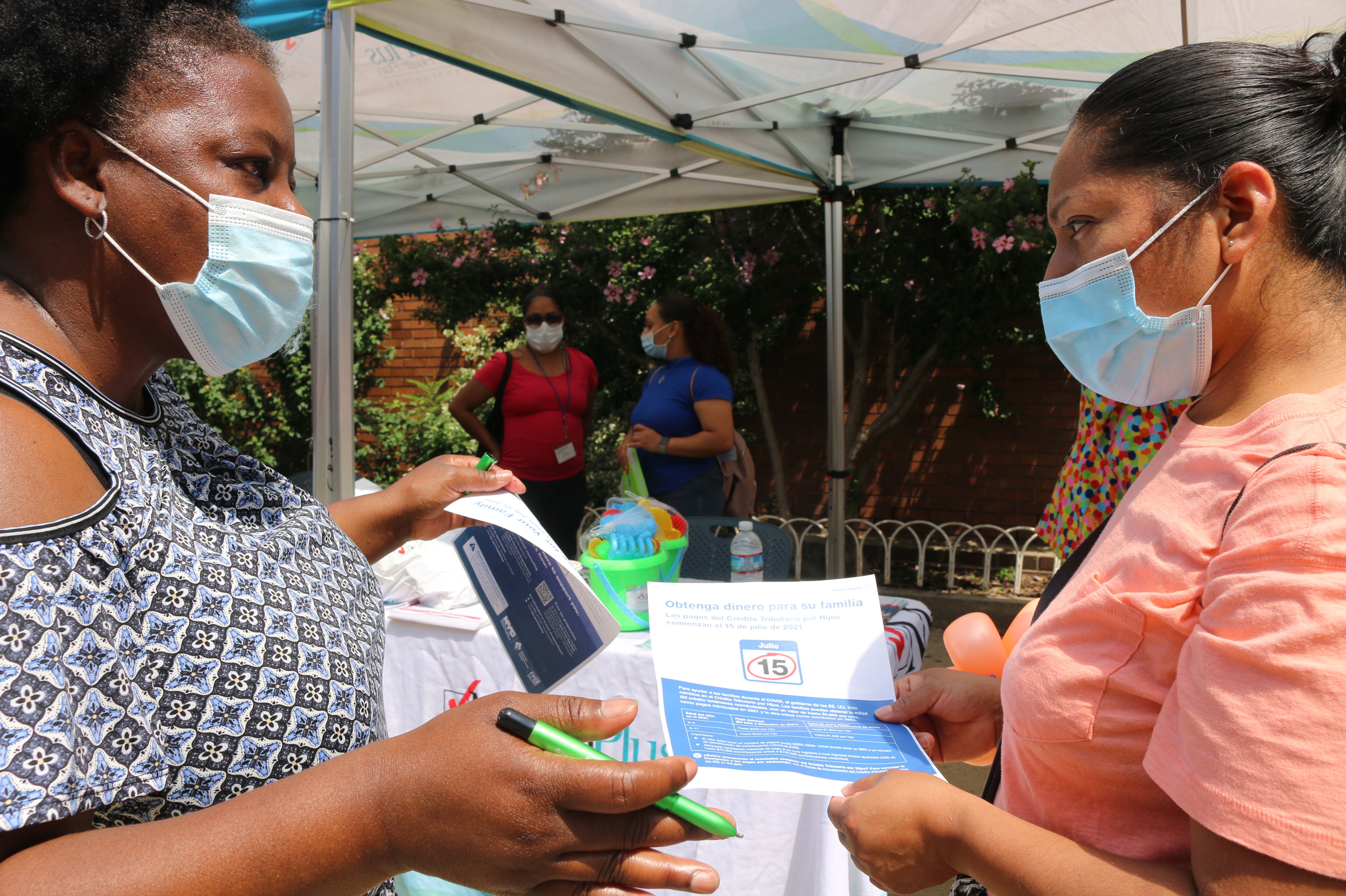 Two middle-aged women of color speak to one another in front of outdoor event tents while holding informational pamphelts.