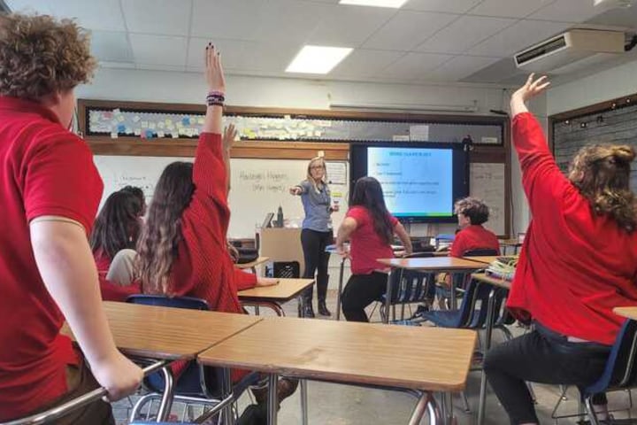 A teacher at the front of a classroom points to students in red shirts who have their hands raised to volunteer for an assignment.