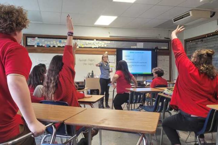 A teacher at the front of a classroom points to students in red shirts who have their hands raised to volunteer for an assignment.