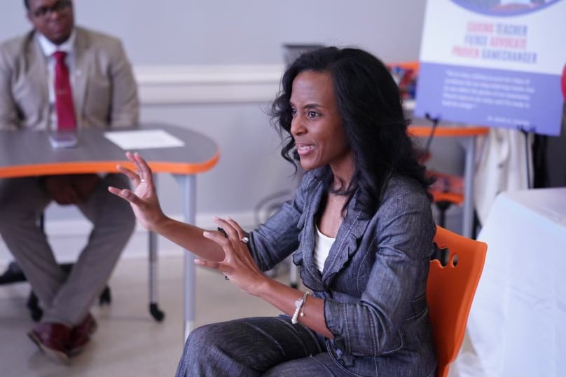 A woman in a charcoal suit sits in an orange chair, explaining a topic she appears to be illustrating with her hands.