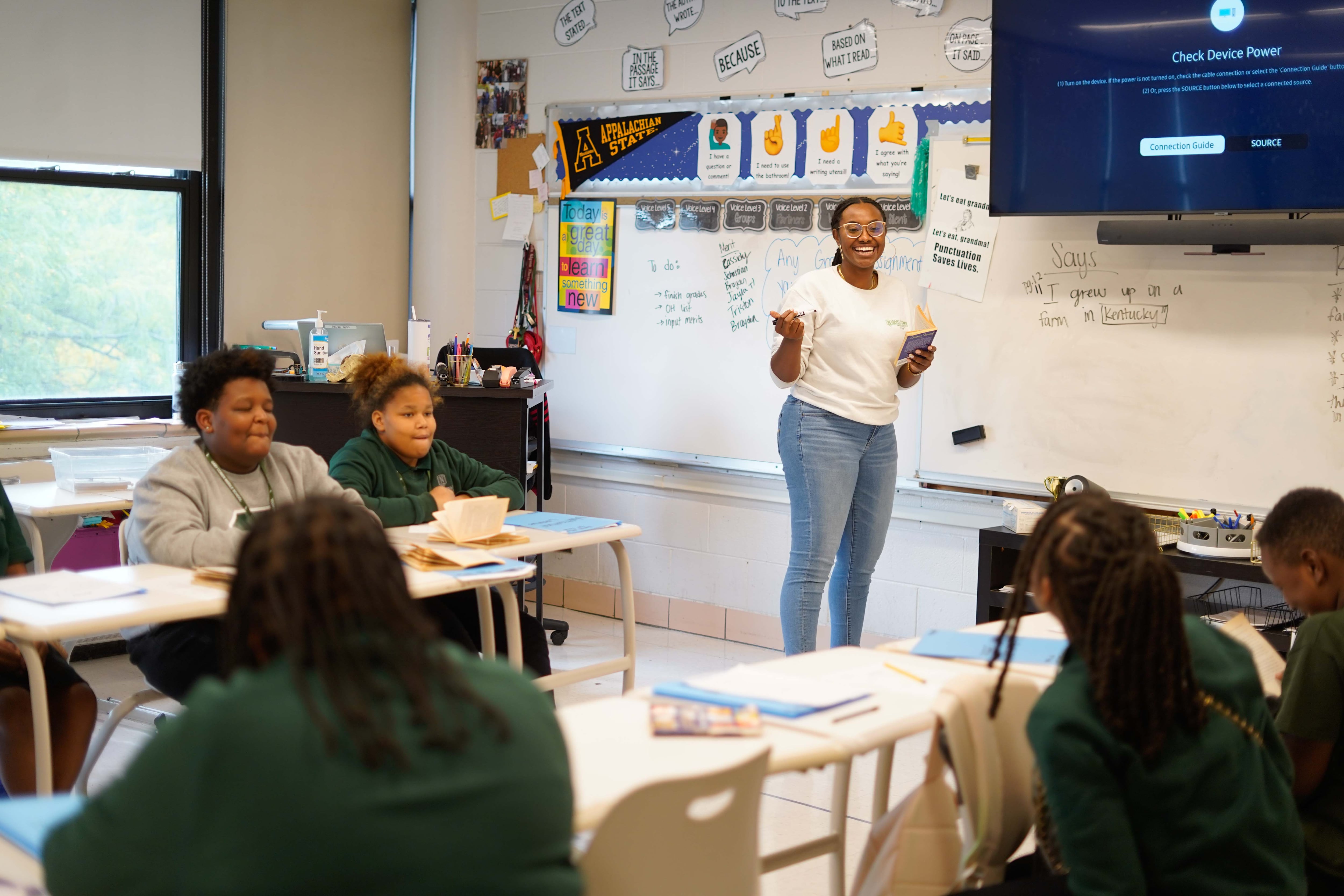 A teacher stands in front of students sitting in desks. There's a whiteboard and different posters in the background.