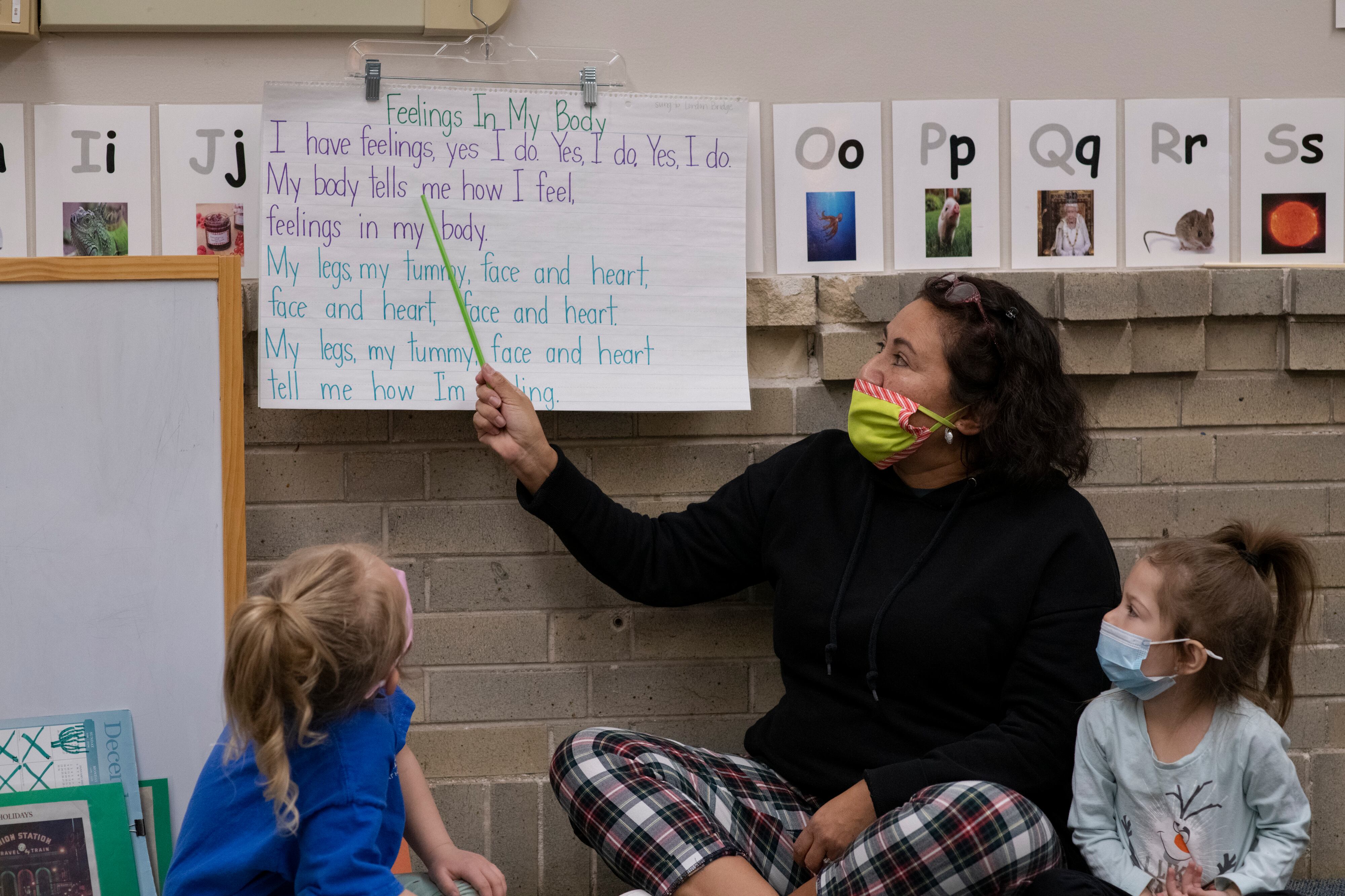 A preschool teacher points to song lyrics written on a big piece of paper as two young students look on. The teacher is wearing a face mask.