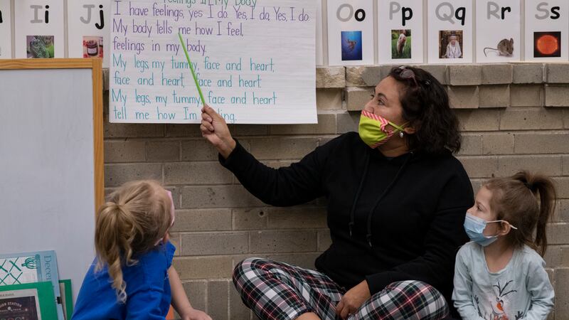 A preschool teacher points to song lyrics written on a big piece of paper as two young students look on. The teacher is wearing a face mask.