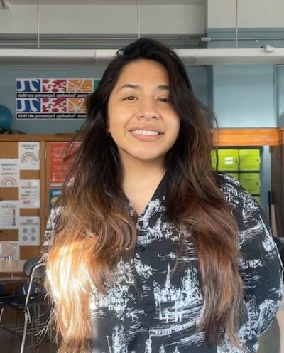 A woman with long brown hair and wearing a black and white shirt smiles while posing for a photo with a classroom in the background.