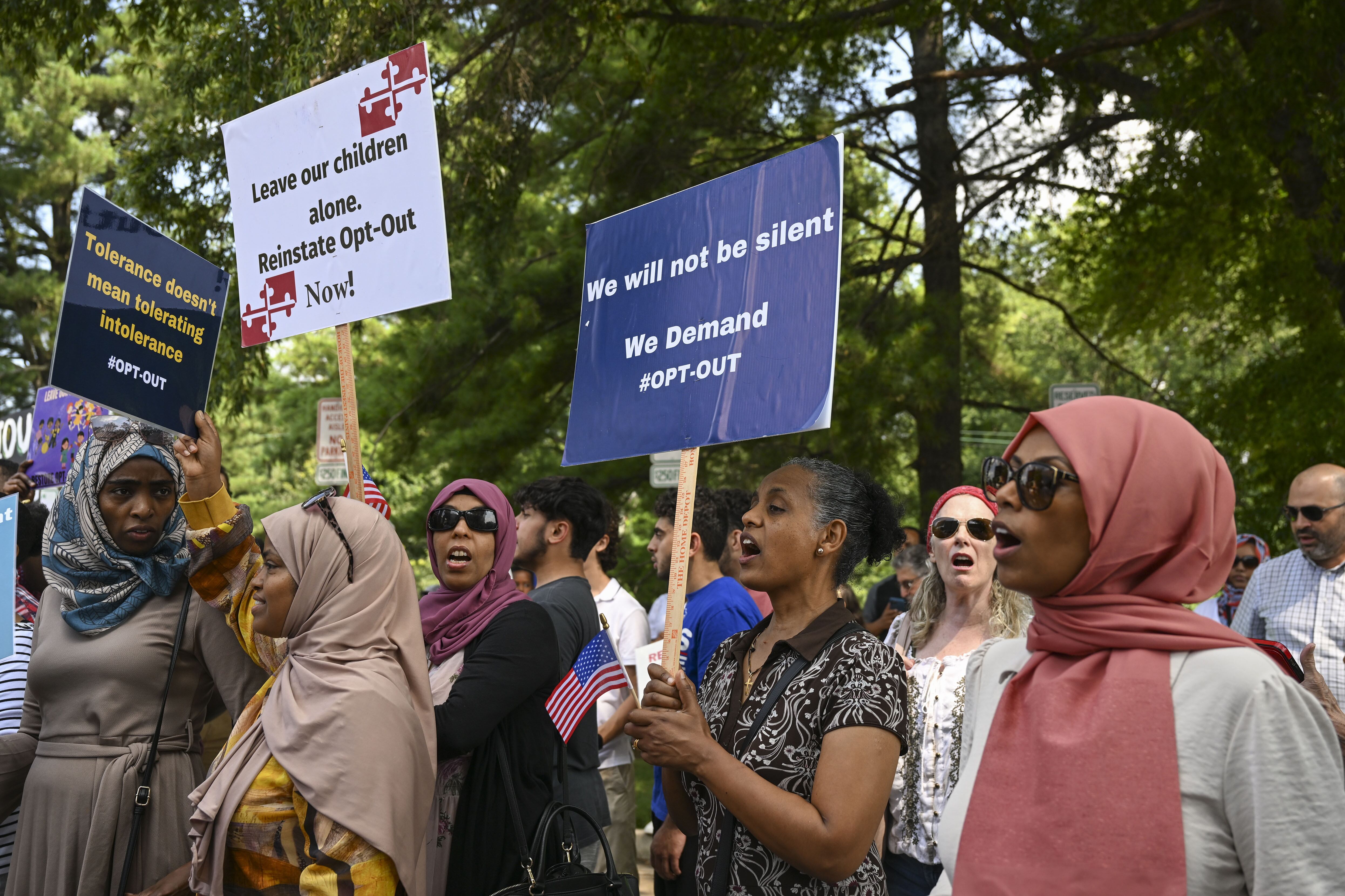 A group of women stand outside in front of green trees during a protest. Some of the women are holding signs.