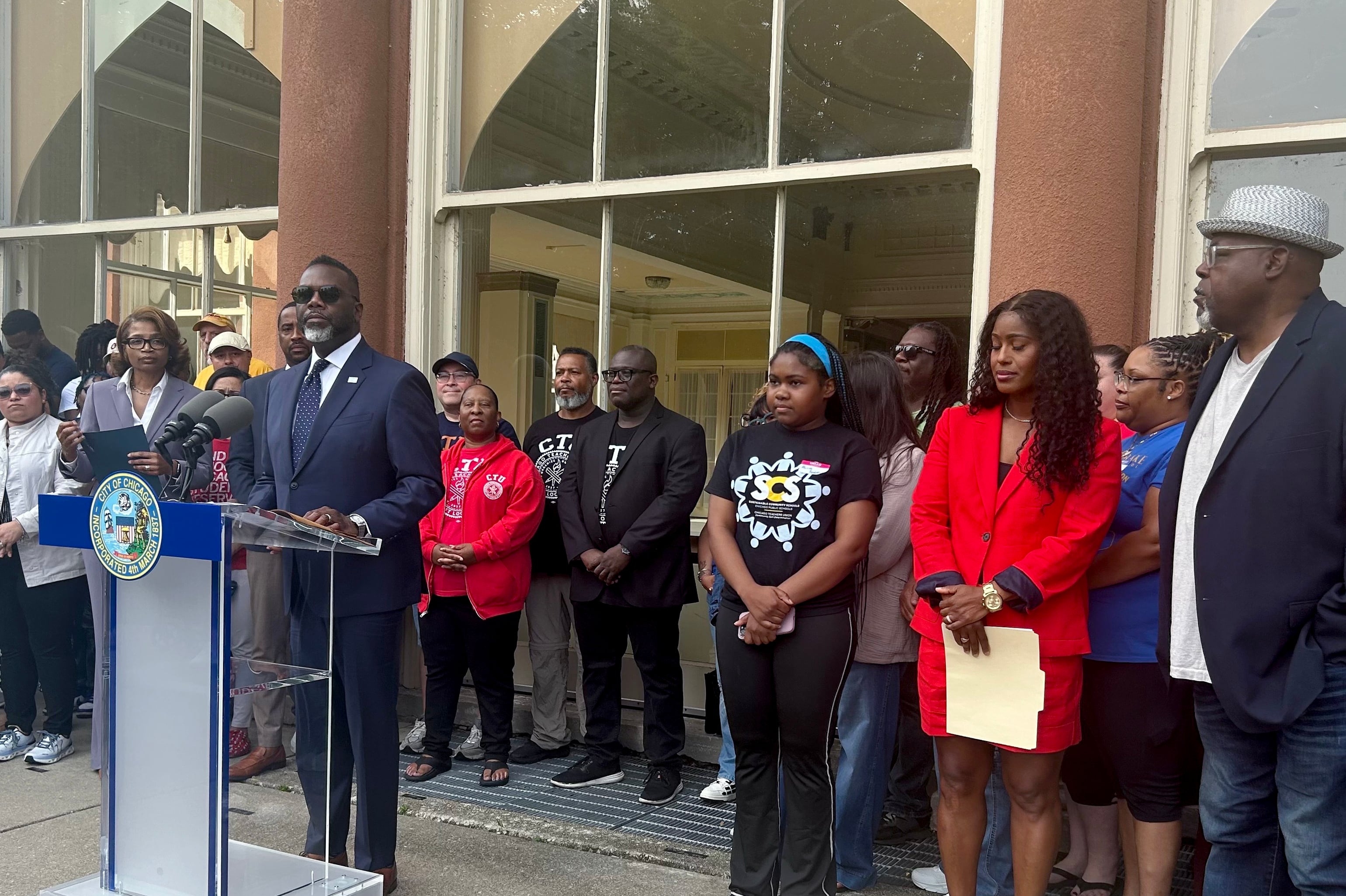 A group of adults stand outside of a large building while one person speaks from a podium.