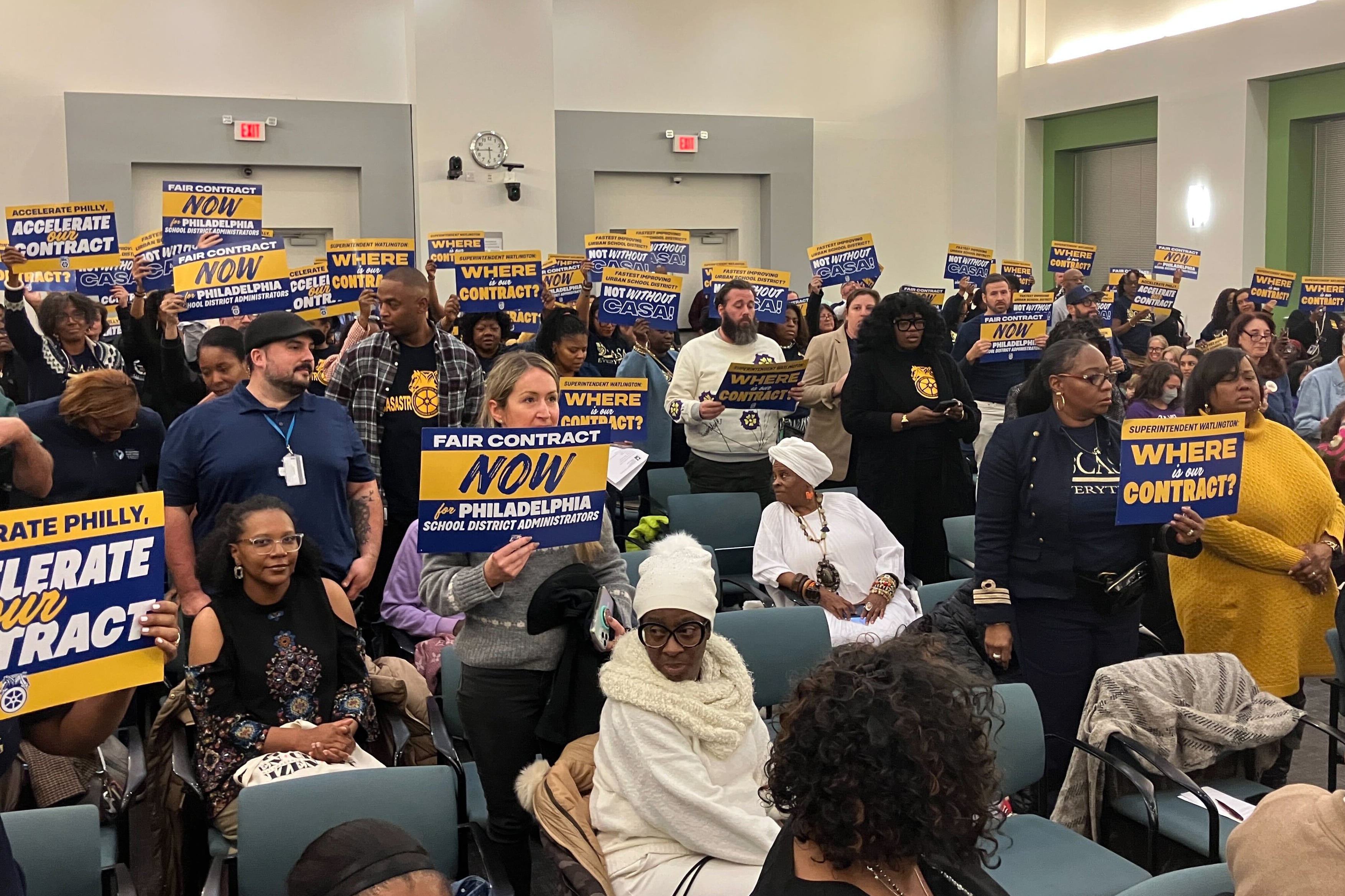 A photograph of a crowded room with adults standing and some sitting with many of them holding signs.