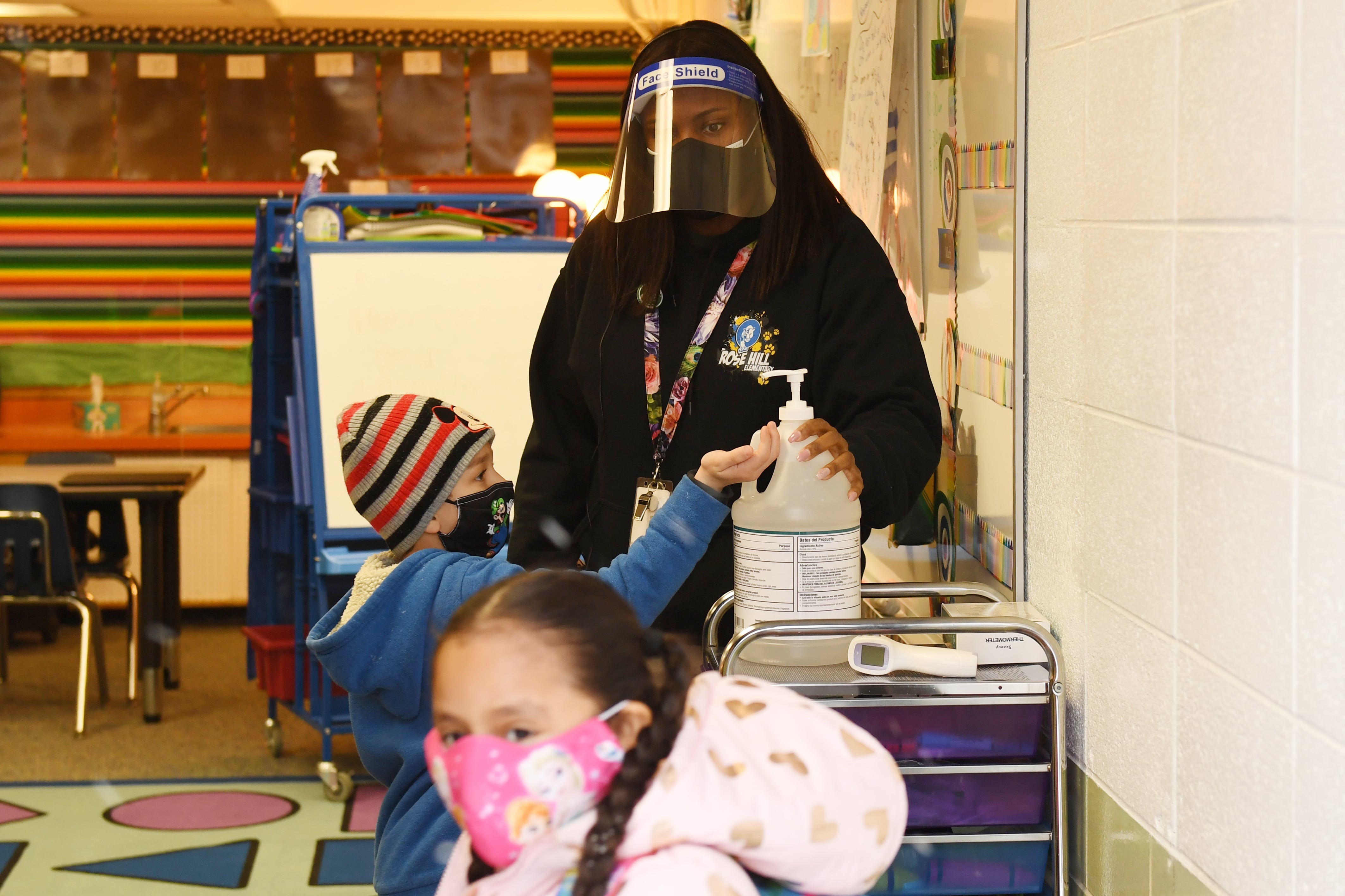 A teacher wearing a mask and face shield helps masked students use hand sanitizer before starting class in an elementary classroom.