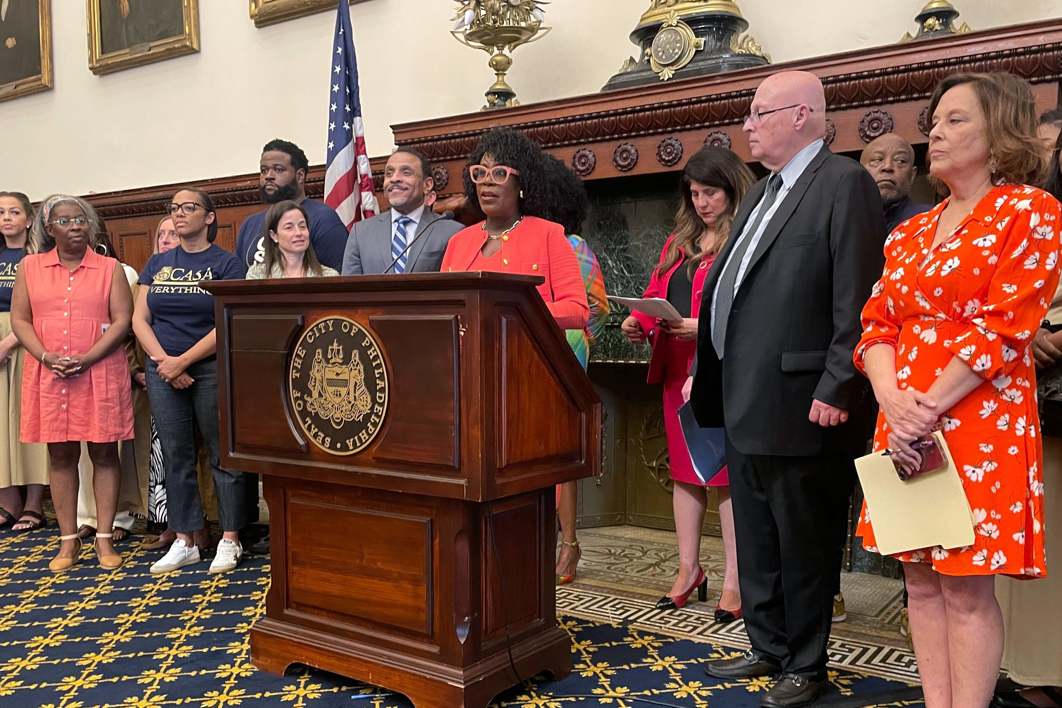 A photograph of a Black woman in an orange suit and glasses speaks from behind a wooden podium with a group of people standing around her in a large meeting room.