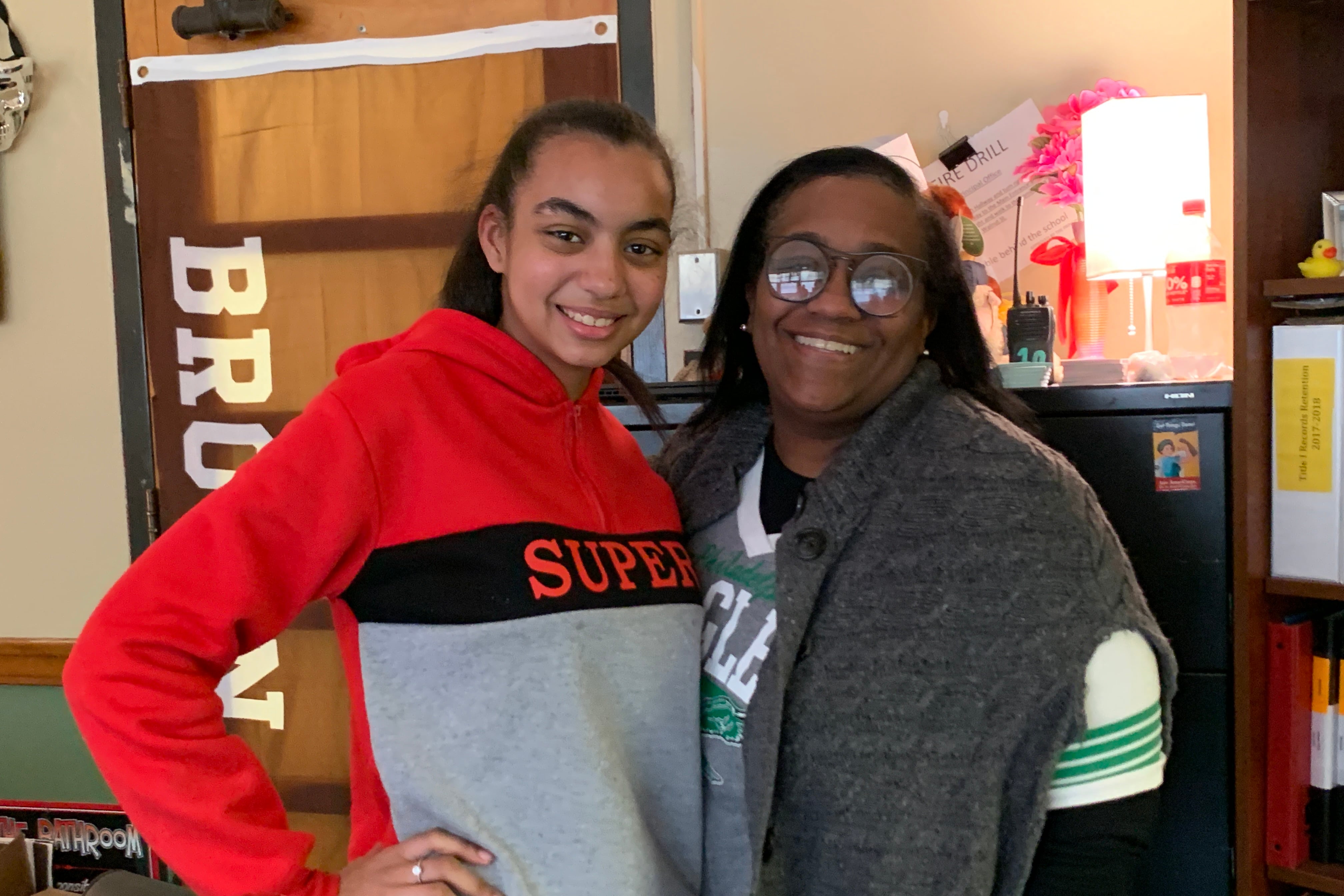 A girl in a shirt that says “Super” stands next to a woman with black hair wearing glasses and a gray sweater over her shoulders.