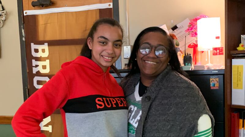 A girl in a shirt that says “Super” stands next to a woman with black hair wearing glasses and a gray sweater over her shoulders. 