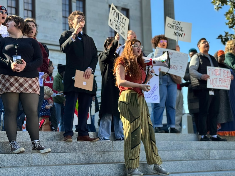 A large crowd of people, many are holding signs, protest outside on a sunny day.