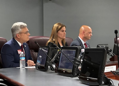 Three people dressed in business clothing sit behind a desk with computer screens and microphones in front of them.