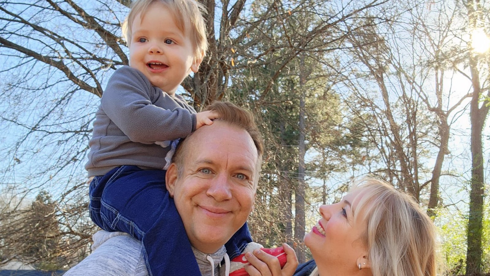 A blond toddler sits on his dad’s shoulders. His mom looks up at him smiling.