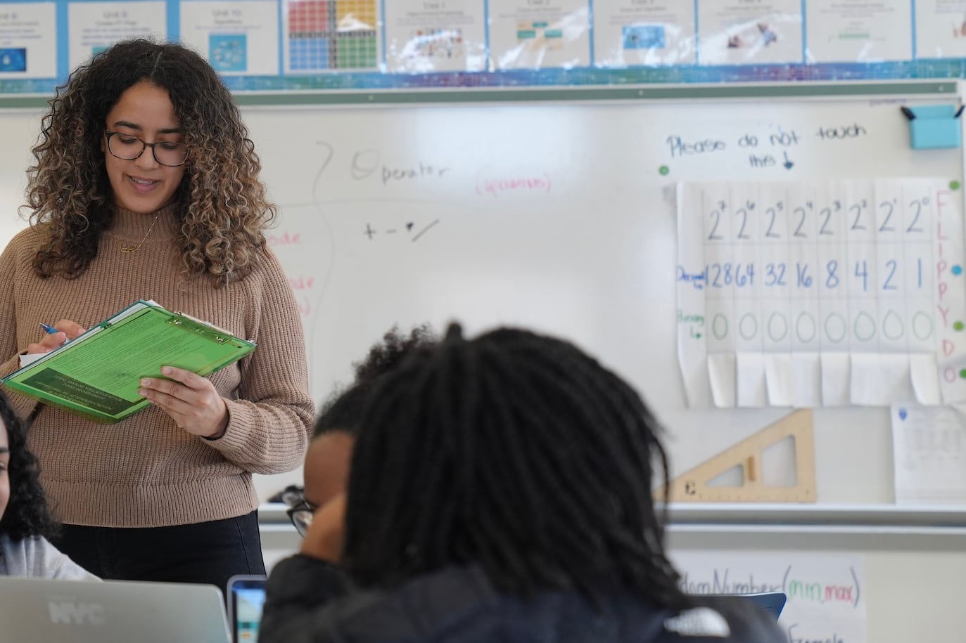 A woman with medium length brown hair and holding a green folder stands in front of two students in a classroom with a whiteboard in the background.
