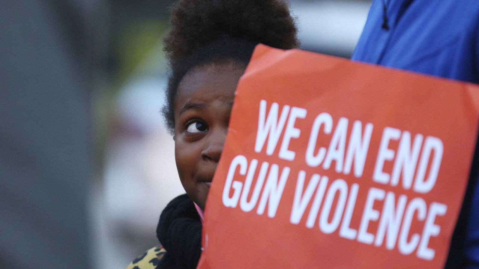 A little girl stands behind a red and white sign that says, “We can end gun violence.” 