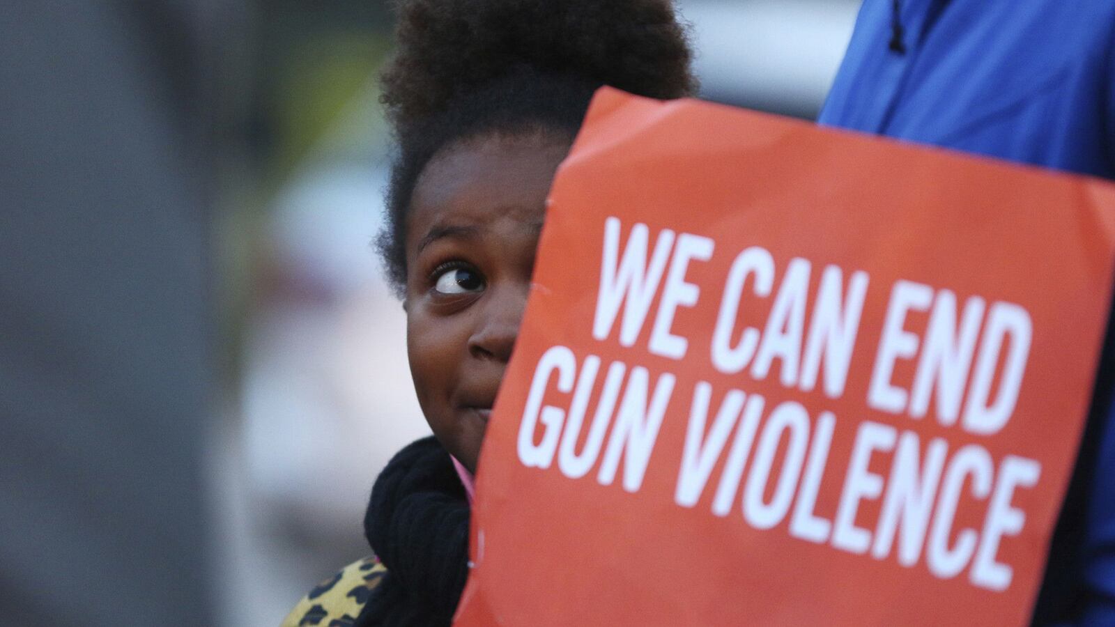 A little girl stands behind a red and white sign that says, “We can end gun violence.”