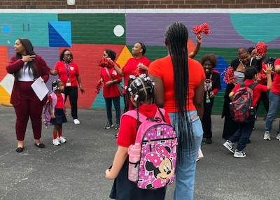 A photograph of a large group of elementary students wearing backpacks and some wearing red shirts while adults cheer them on.