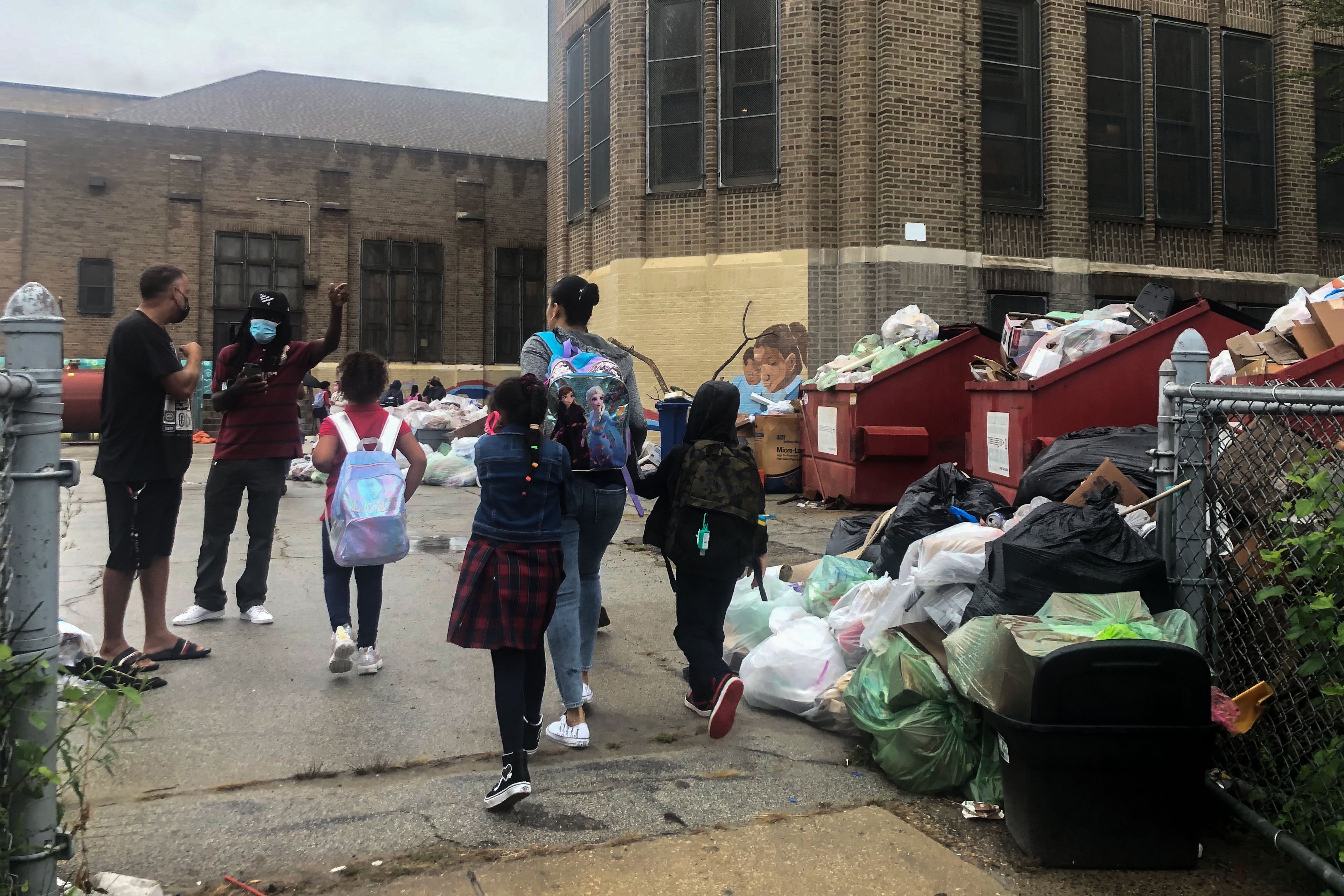 Children and adults were dark clothing walk past clear and brown colored trash bags.