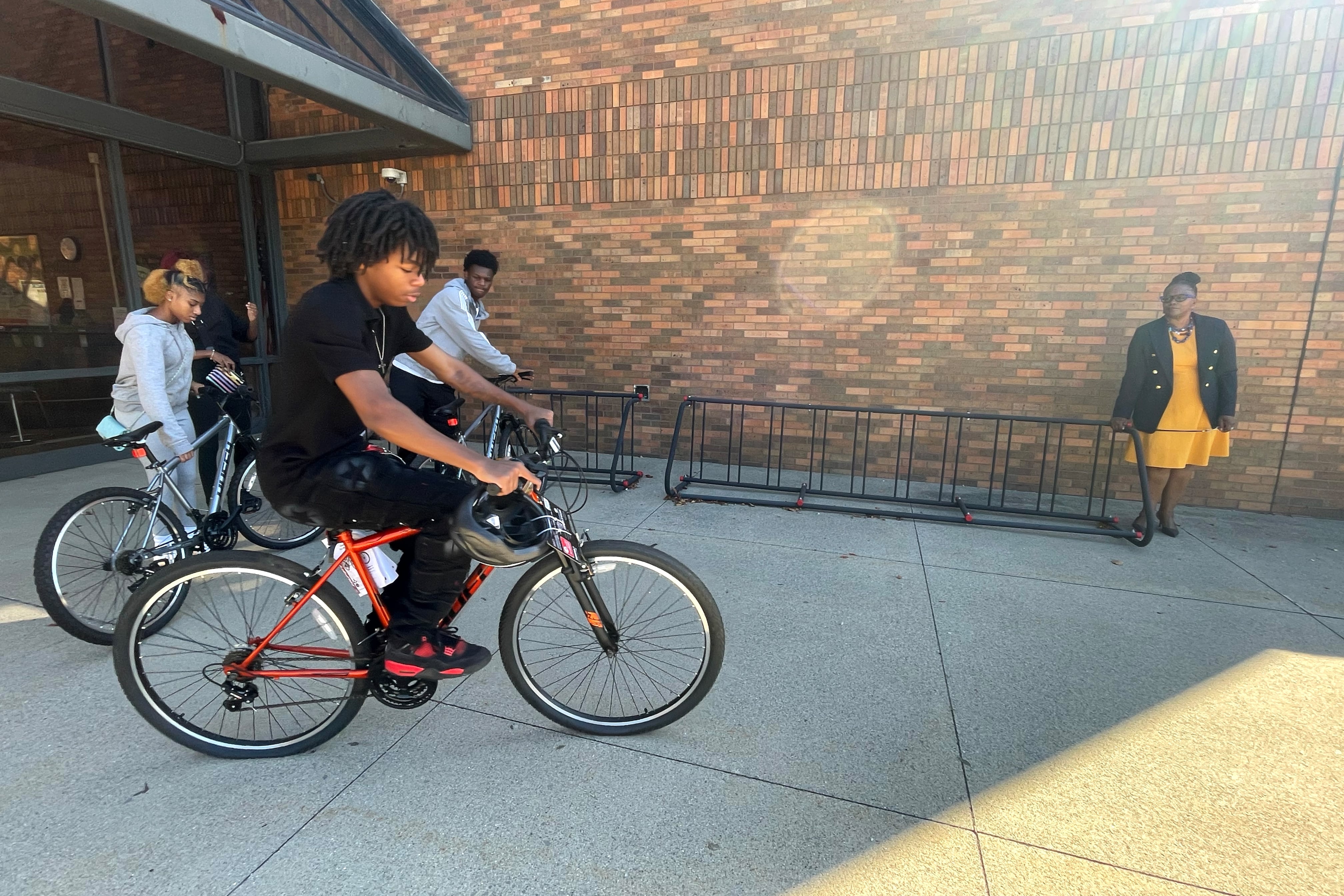 Students on bikes are in front of a woman in a dark jacket and orange dress.