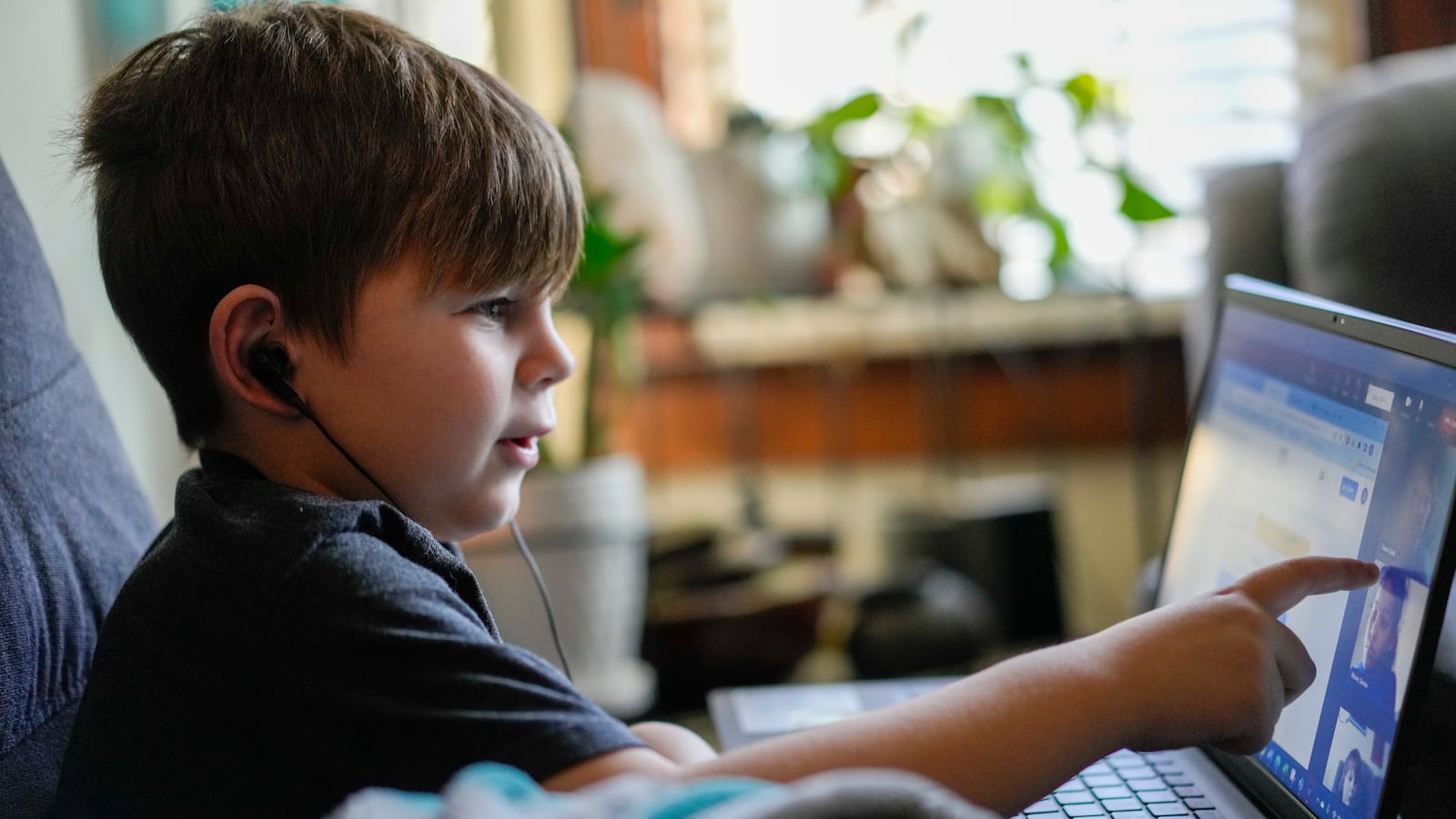 A young boy works on his laptop while sitting on a couch, pointing at the screen while wearing headphones.