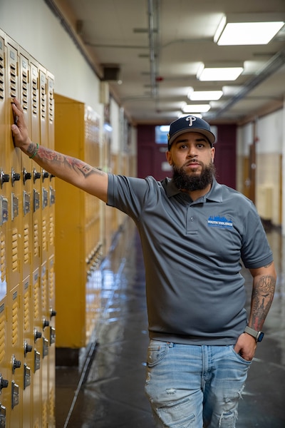 A man wearing a baseball cap and blue shirt poses for a photograph in a hallway with yellow lockers