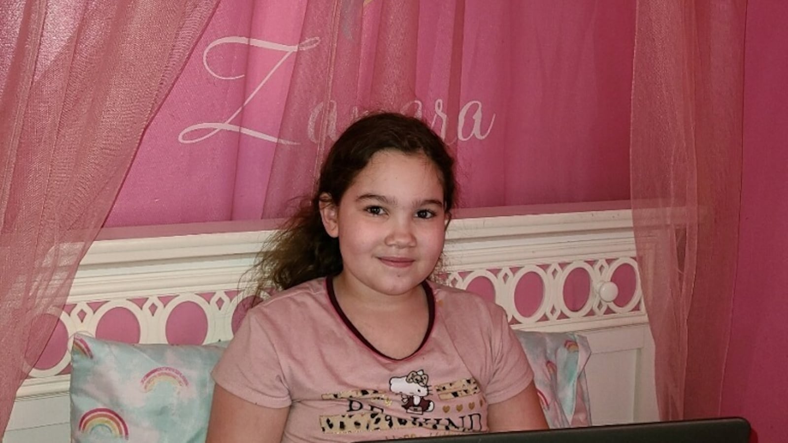 Chicago student Zamara sits with her laptop on her bed, she has pink curtains and a blue bedspread.