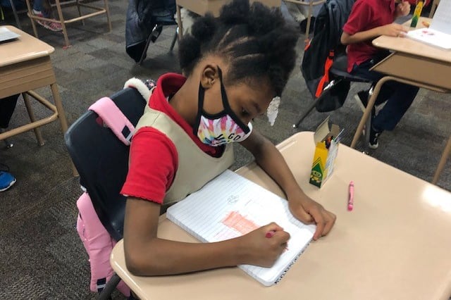 A student wearing a face mask sits at a desk and draws in a notebook with crayons on the first day IPS returned to classrooms during the pandemic.