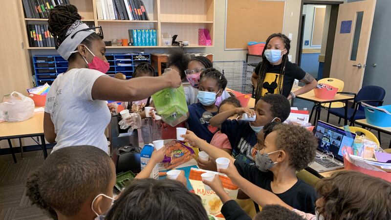 Children in a classroom wearing face masks hold out white paper cups to another student, who is pouring a green smoothie from a blender.