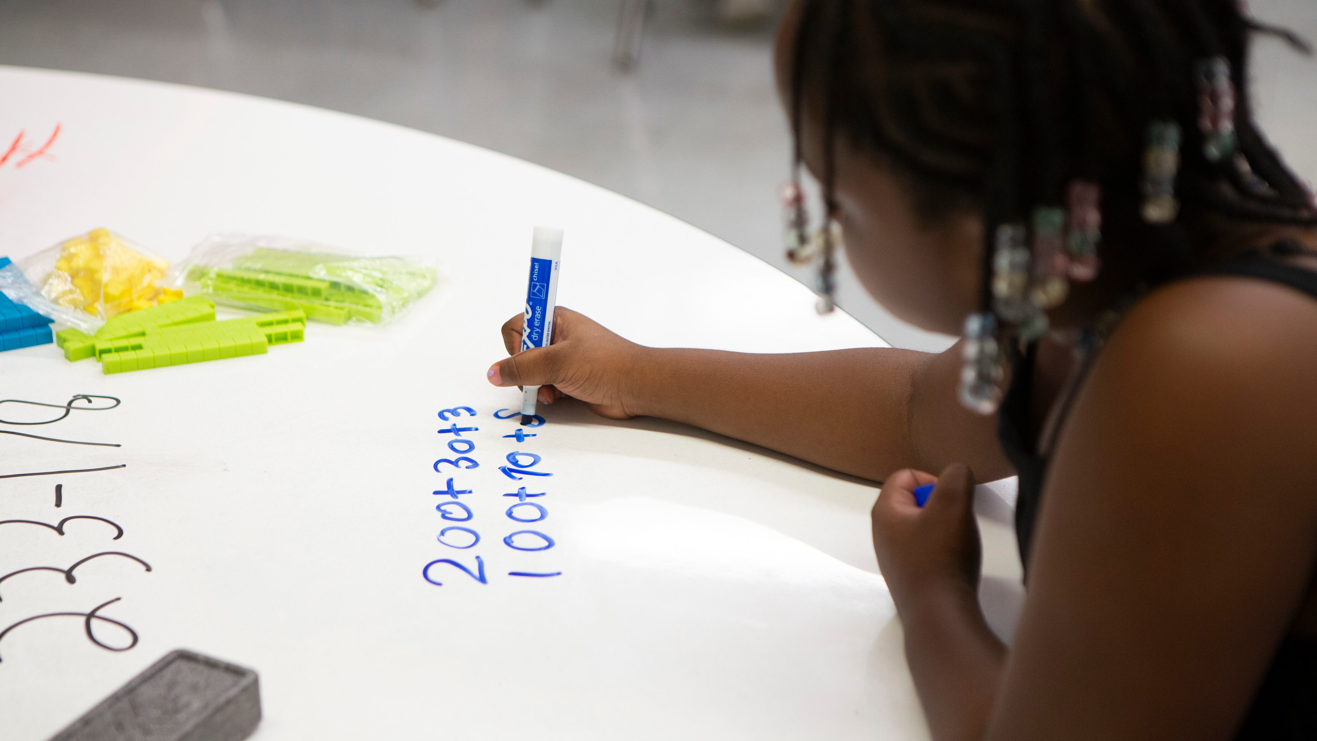 A young girl holds a blue marker in her hand as she sits at a desk and writes an equation on poster paper.