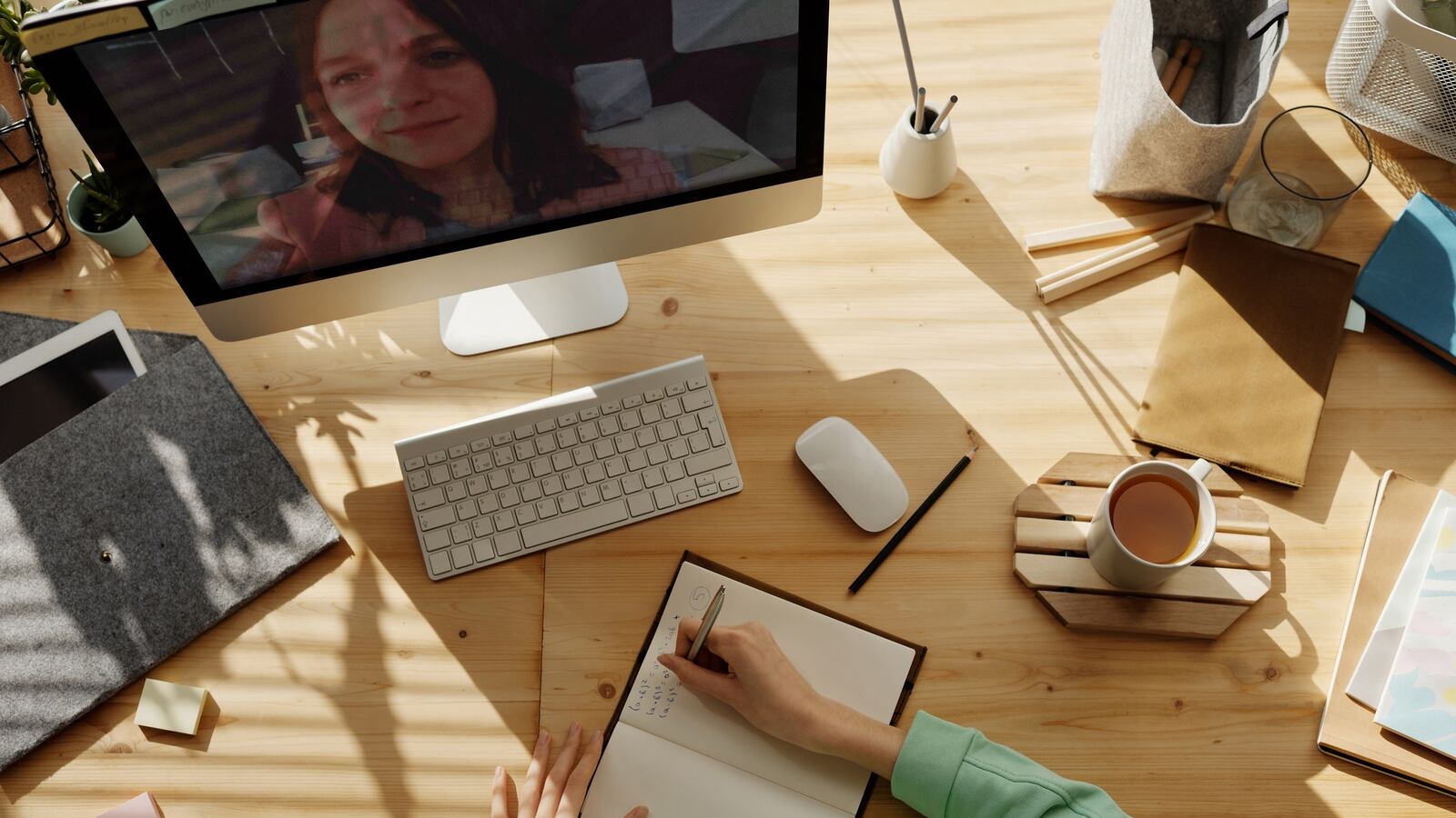A teacher’s face is on a computer screen, and a student is sitting at a desk with a notebook open, pen in hand, taking notes. Light is filtering in on the table through blinds, and a cup of tea is on the desk, along with a few other office supplies including pencils.