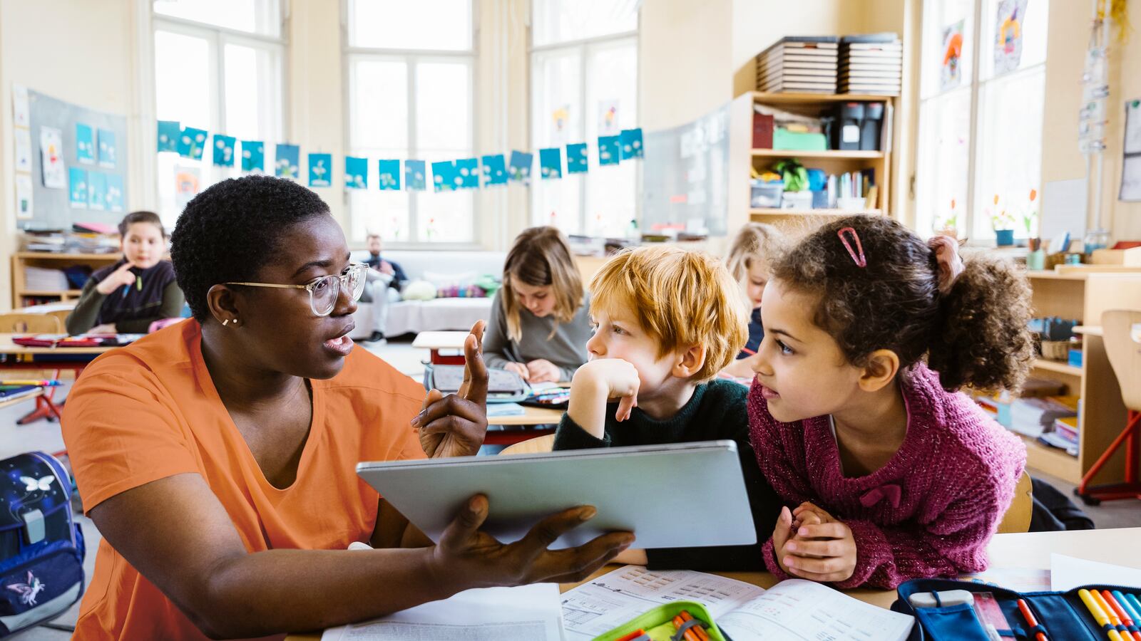 An adult with short dark hair and wearing an orange shirt, holds a book while sitting next to two young students in a classroom with more students sitting at their desks in the background.