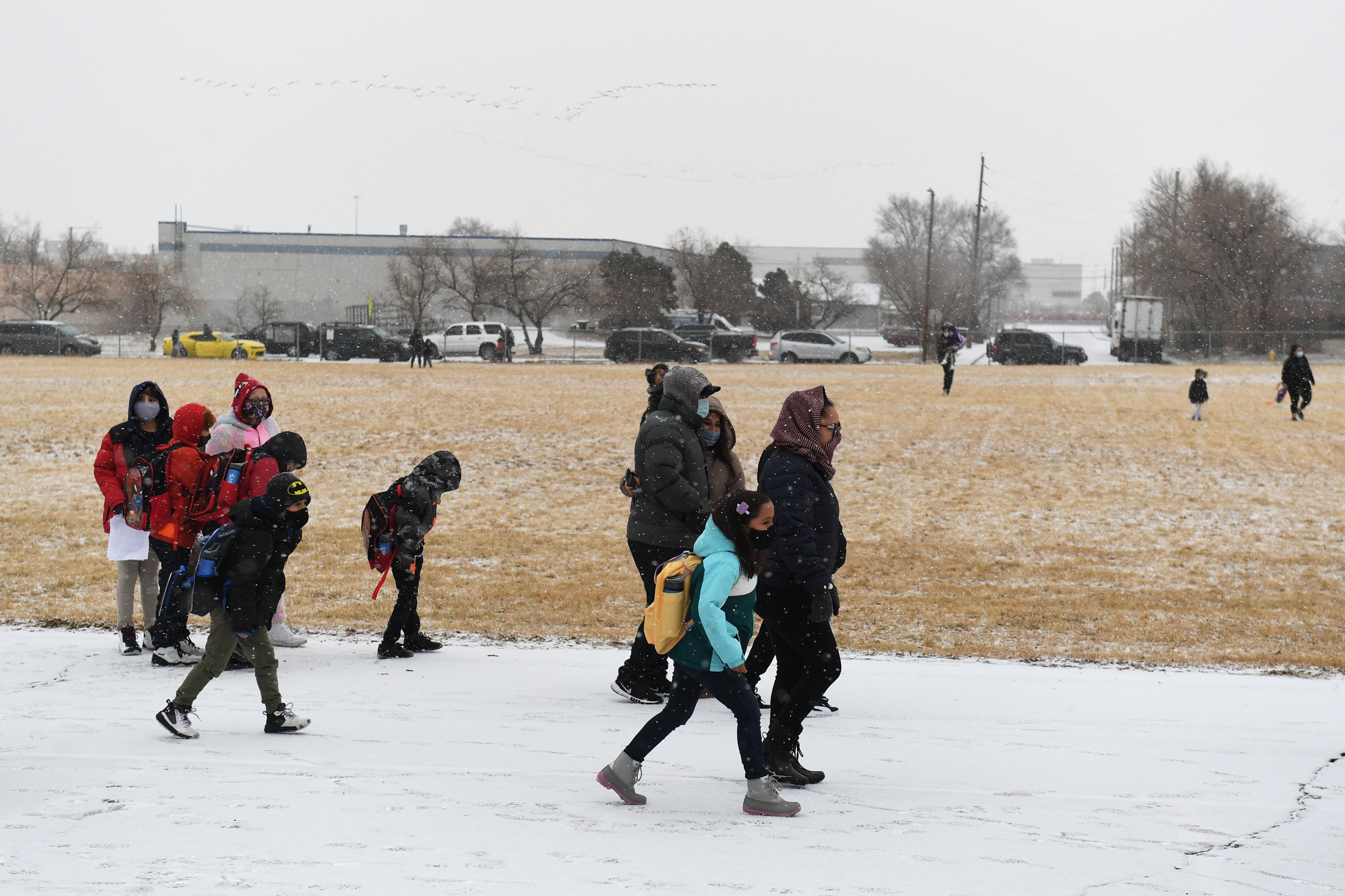 Children and parents wearing masks walk to school across a snowy field.