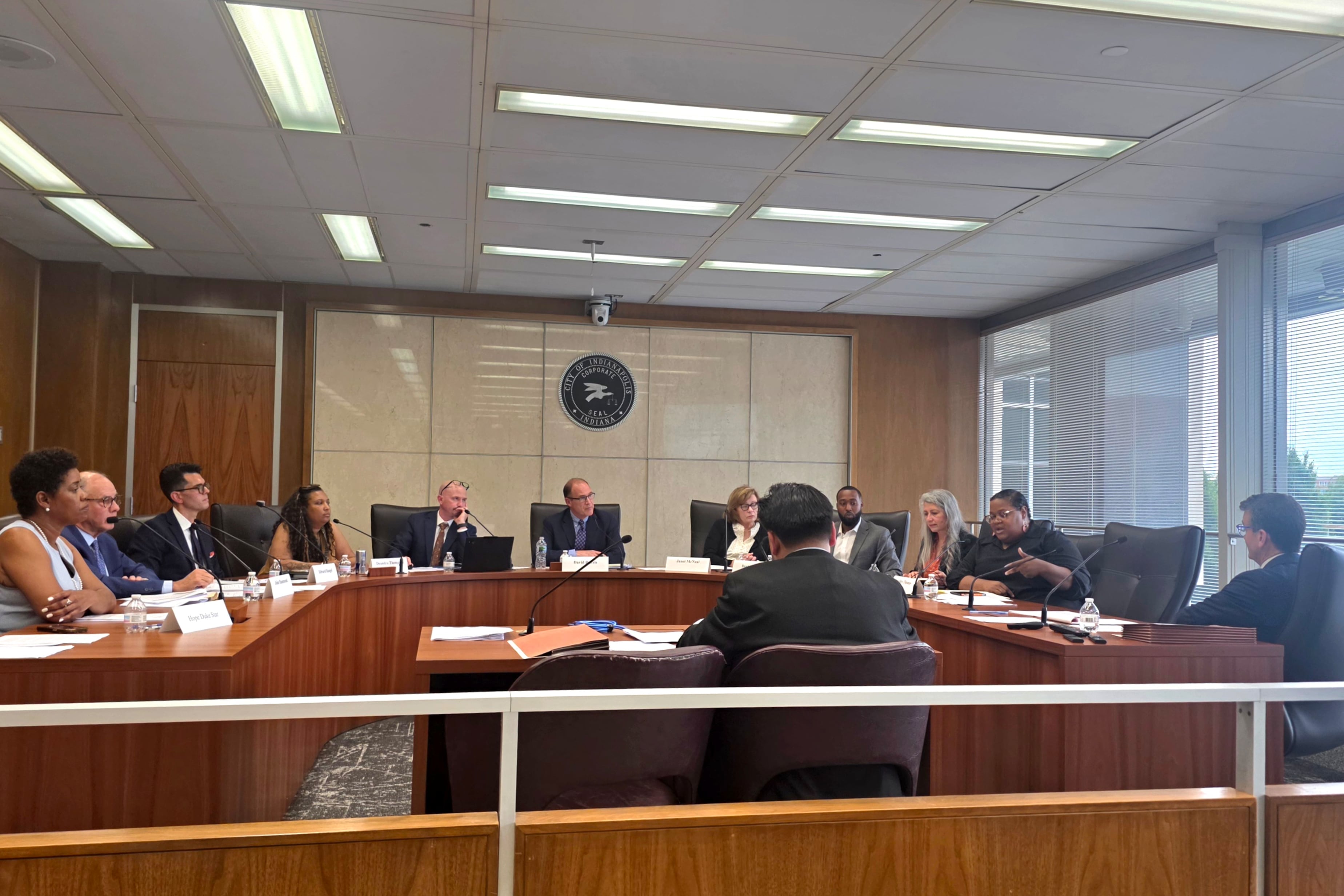 A photograph of a large group of adults in business clothes sitting around a wooden table in a meeting room.