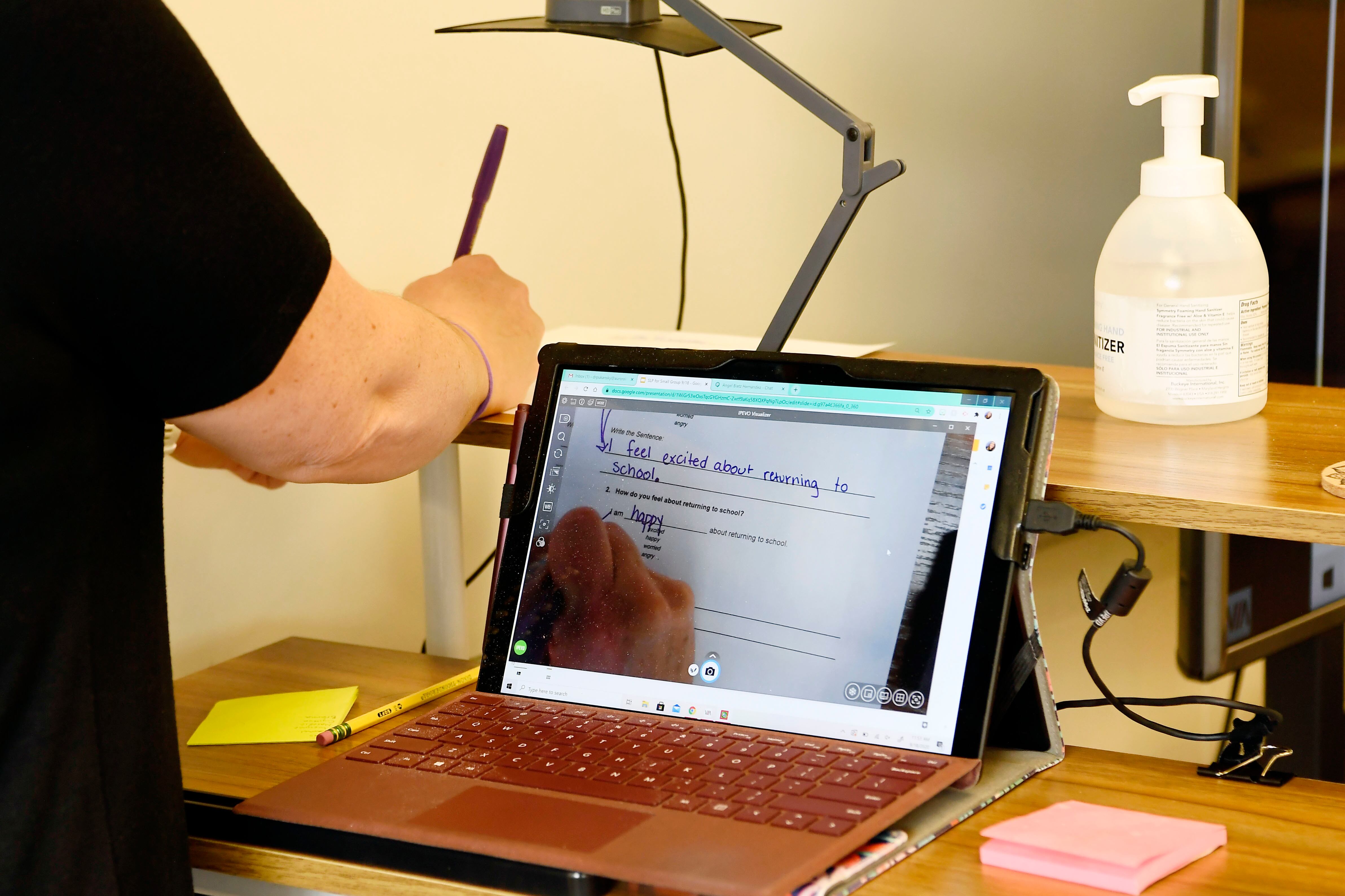 Back view of a standing teacher using an overhead projector that shows her hand writing as she reviews a worksheet.