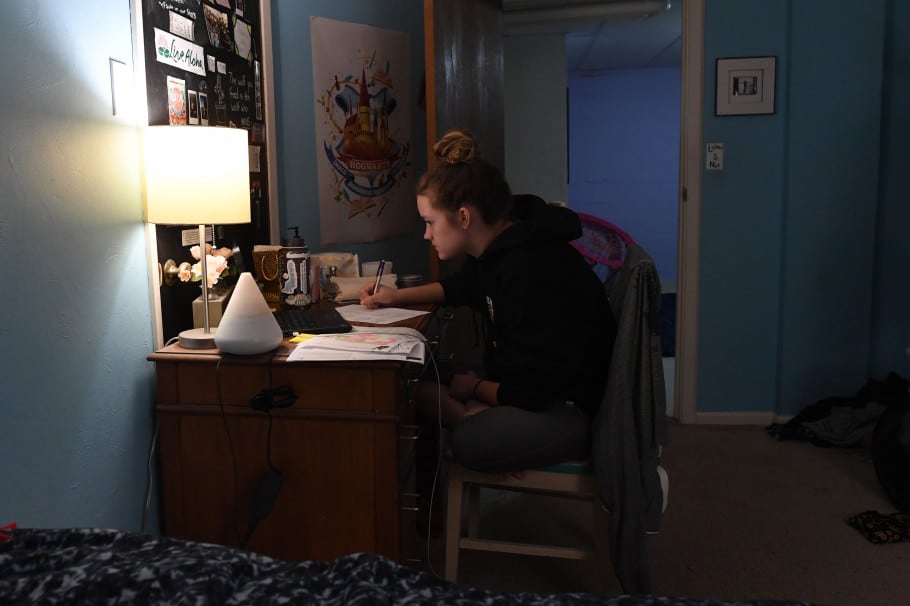A student engages in remote learning from her bedroom, using a lamp to light her work.