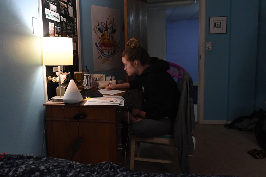 A student engages in remote learning from her bedroom, using a lamp to light her work.