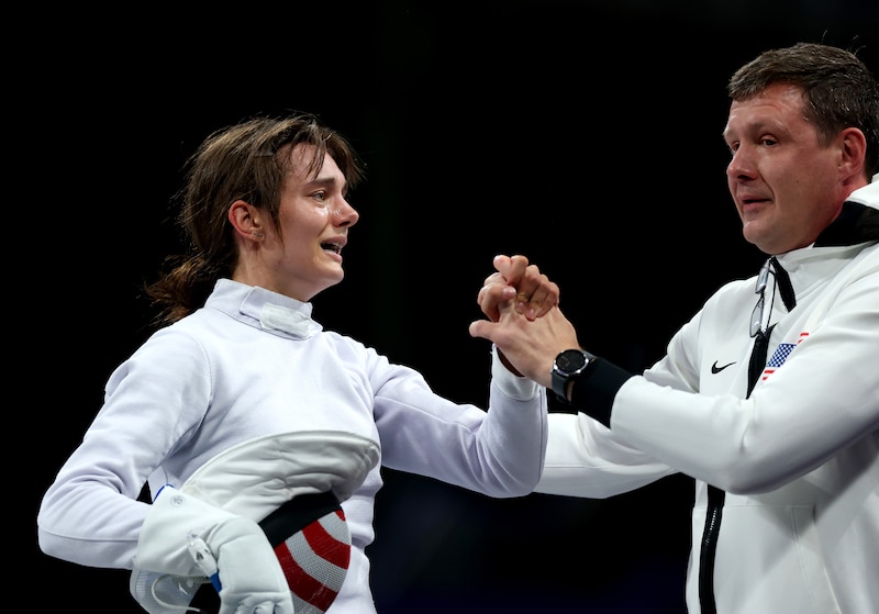 A young woman with dark hair and wearing a fencing uniform grabs the hand of a man wearing all white after winning her match with an all black background.
