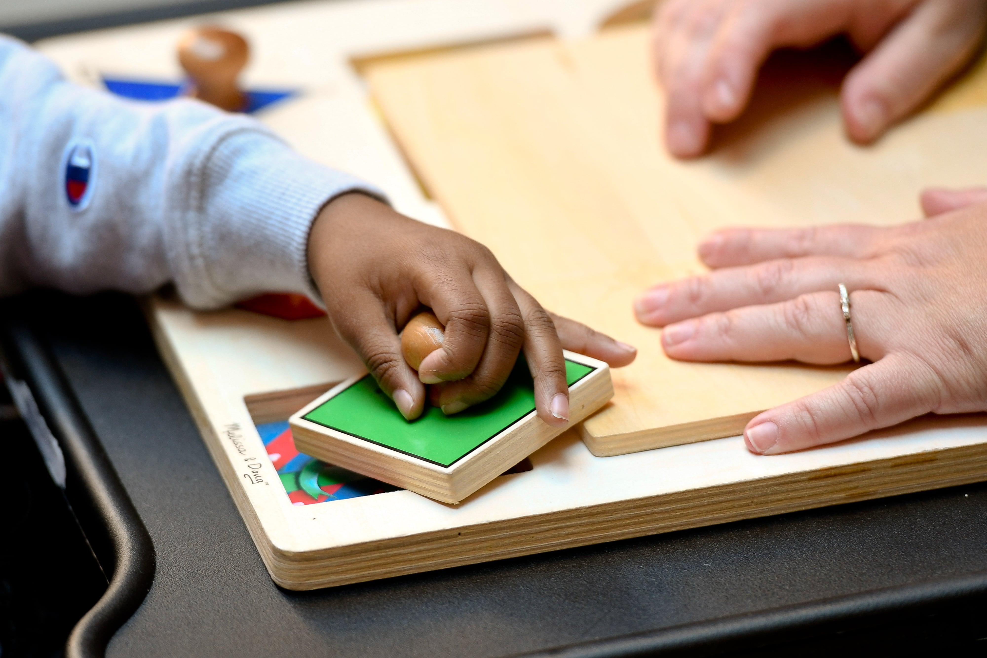 The hand of a young boy works on a small puzzle exercise with assistance from his teacher, whose hands steadies the puzzle.